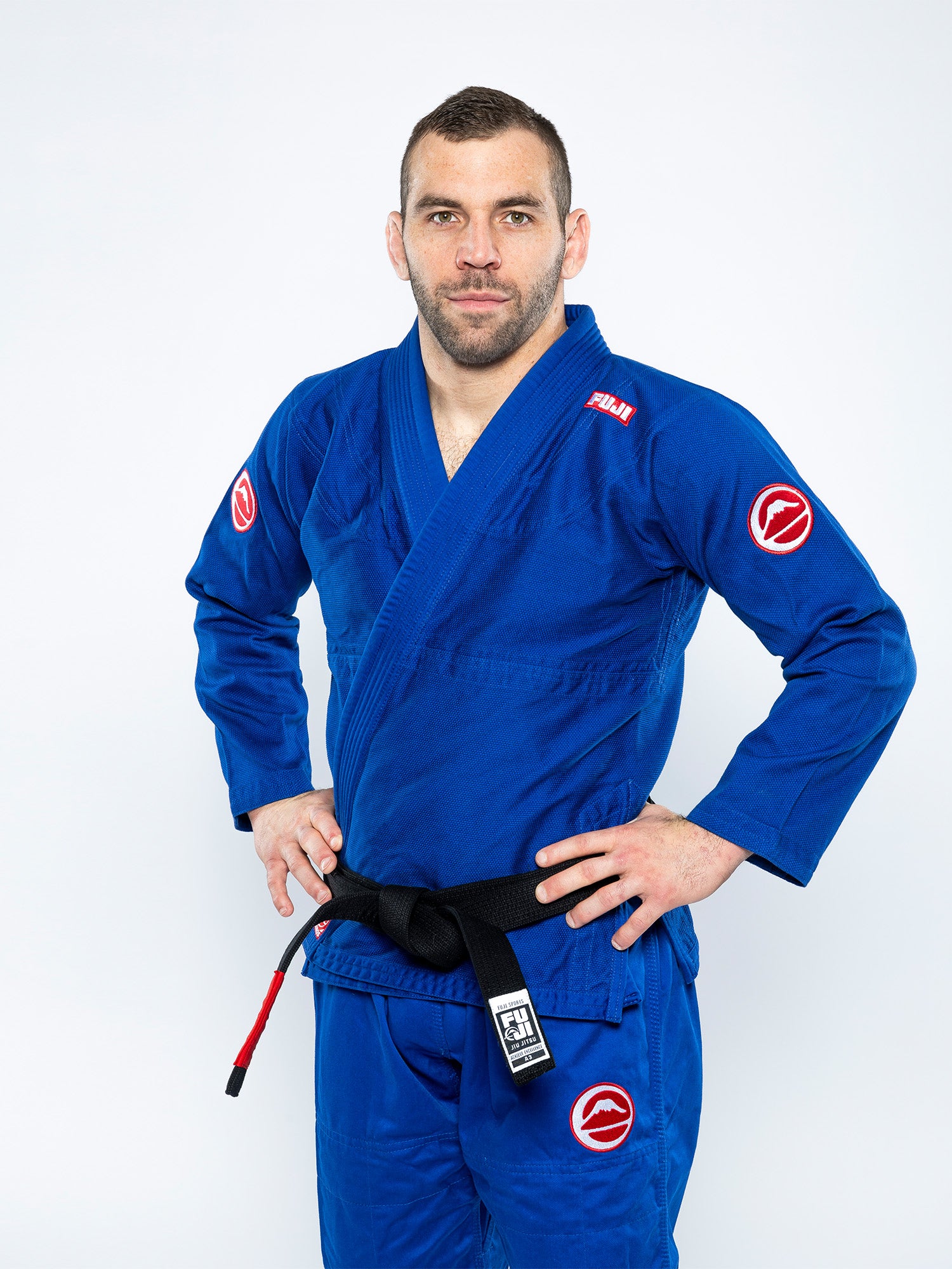 A man confidently stands with his hands on his hips, wearing the Competition BJJ Gi Blue and a black belt, set against a plain white background.