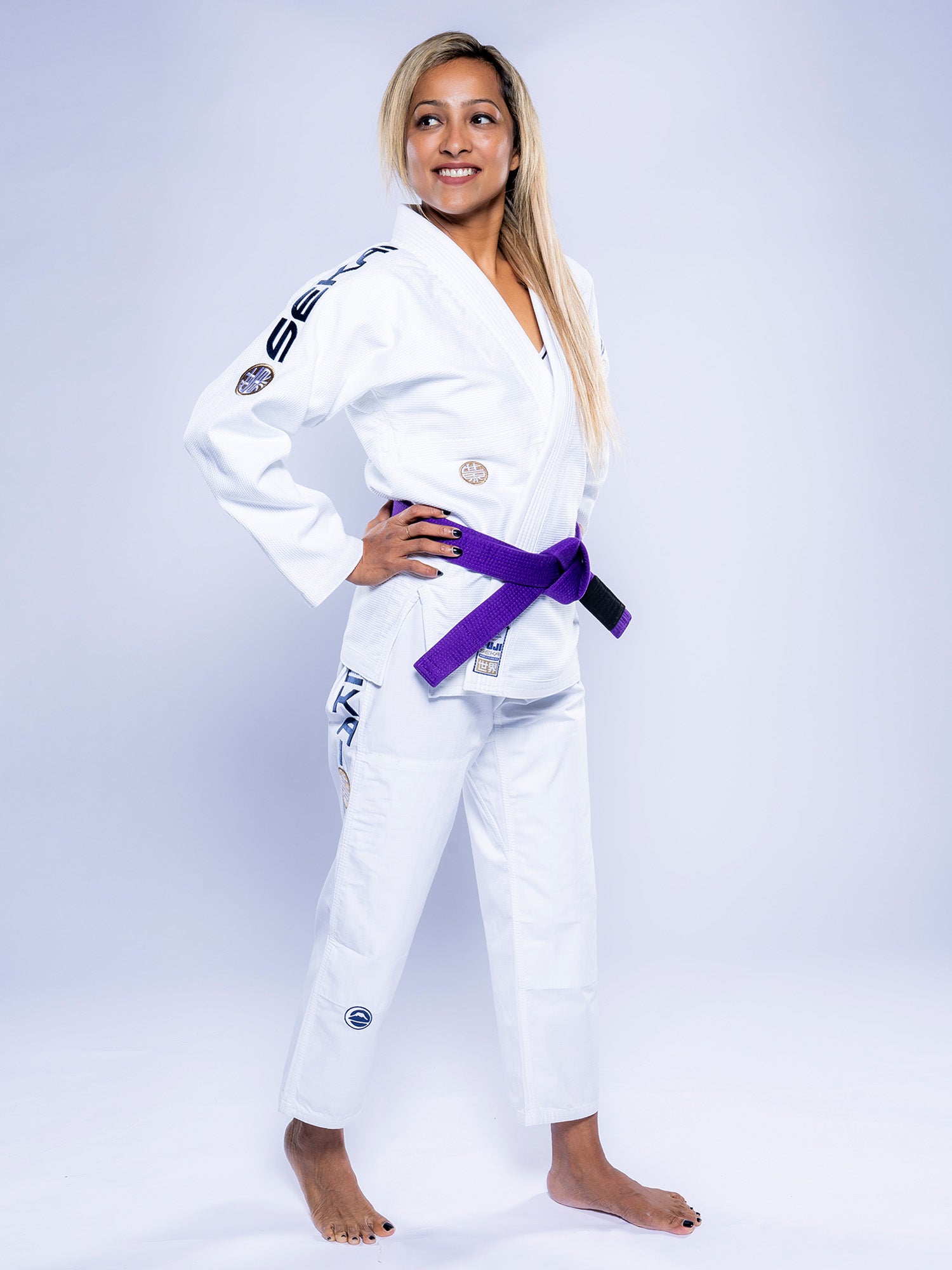 A woman smiles and looks to the side, standing barefoot with hands on her hips in a Womens SEKAI 3.0 BJJ Gi White Navy and a purple belt, against a plain light background.