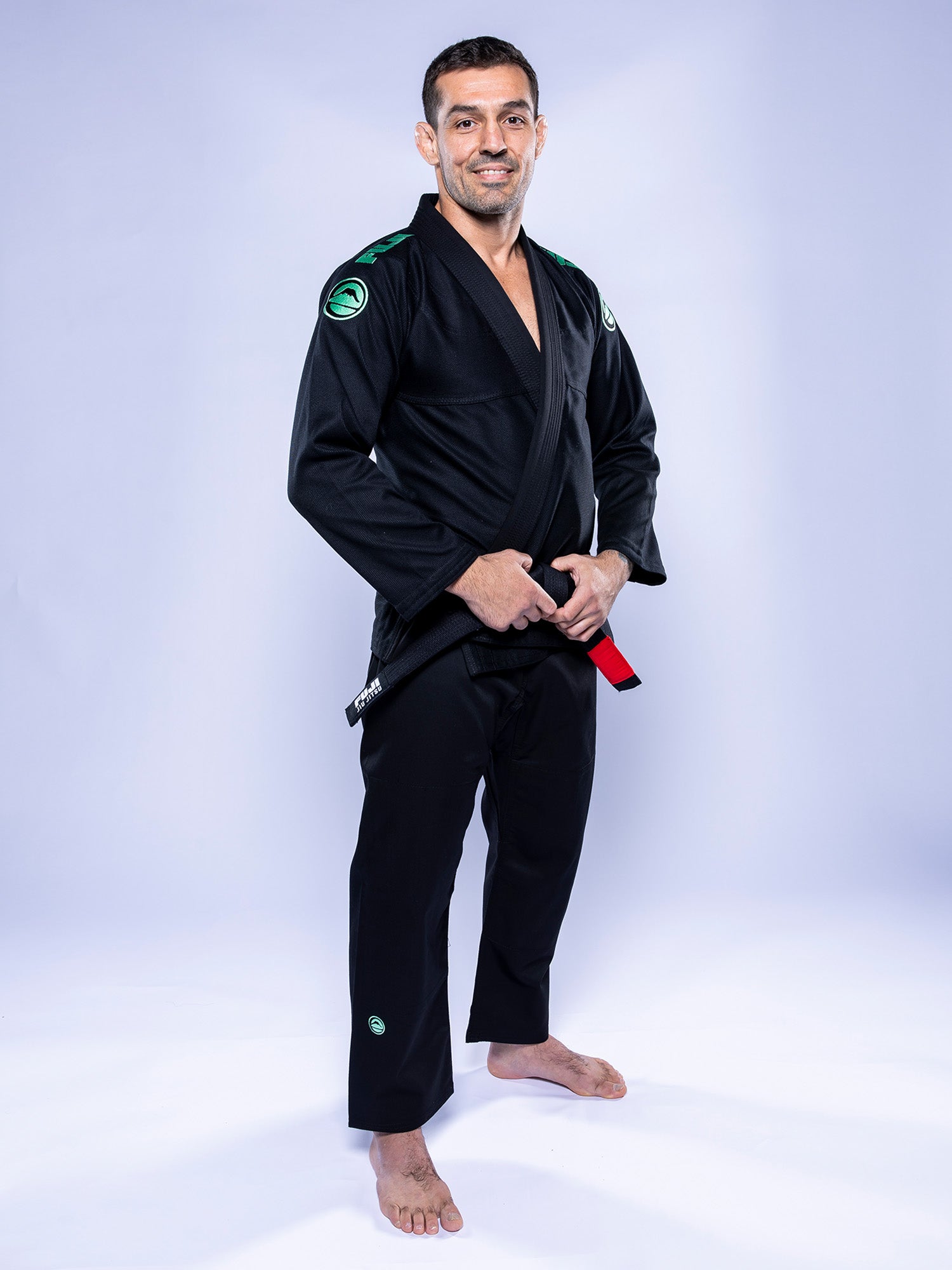 A man wears the Baseline 2.0 BJJ Gi Black Mint, standing barefoot on a white background, smiling and holding a black belt with a red stripe.