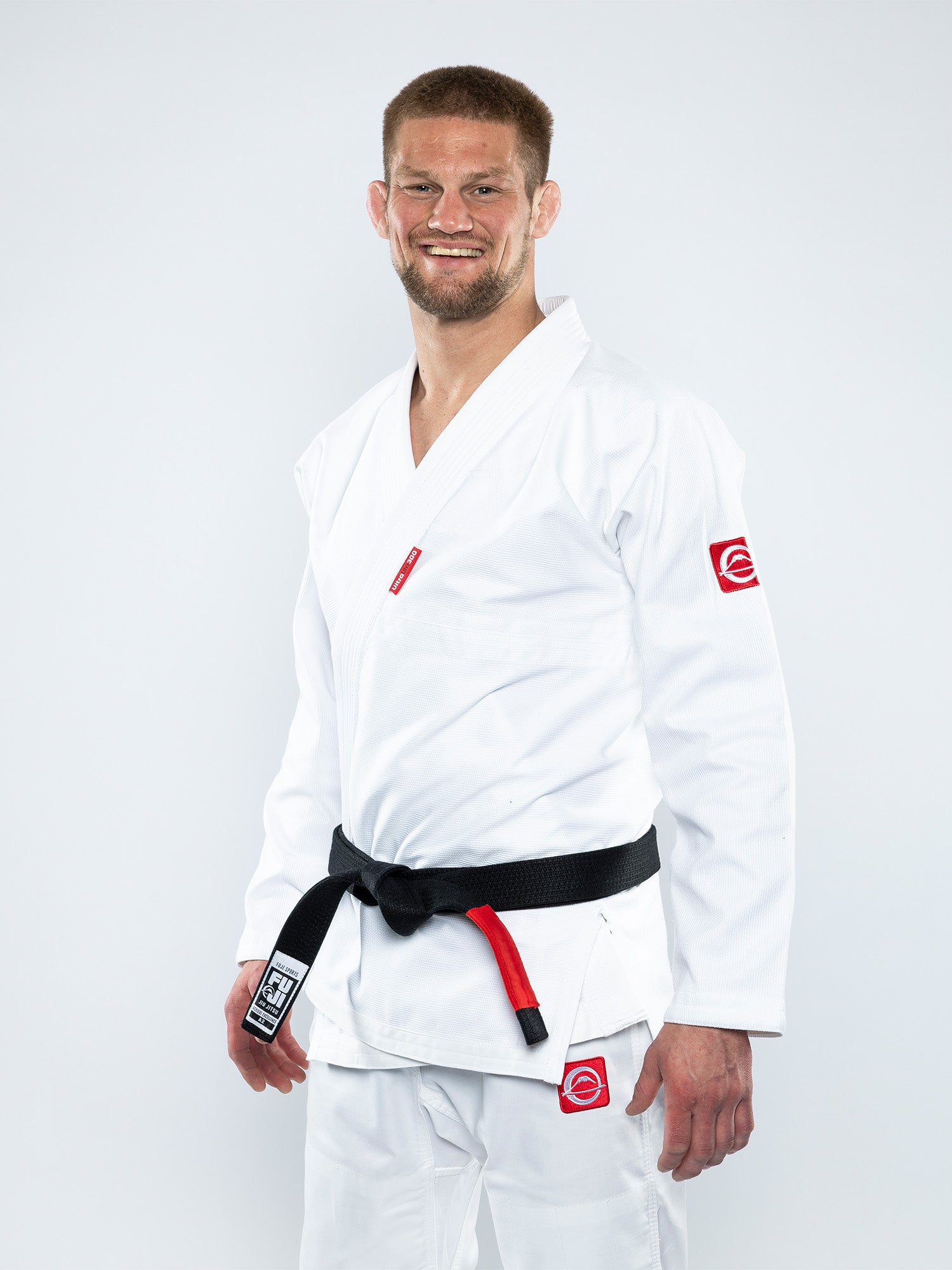 A man wearing the Ultra Lite 300 BJJ Gi White with red patches and a black belt stands smiling against a plain white background.