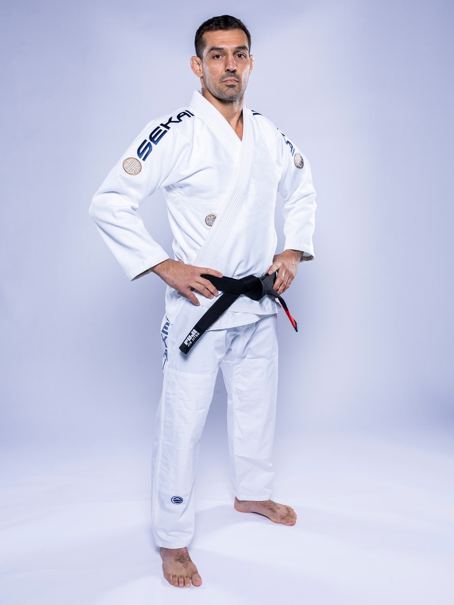 A man stands confidently on a white background wearing the SEKAI 3.0 BJJ Gi White Navy with a black belt, hands on hips, looking straight ahead. The gi features distinctive patches and logos on the sleeves and legs.