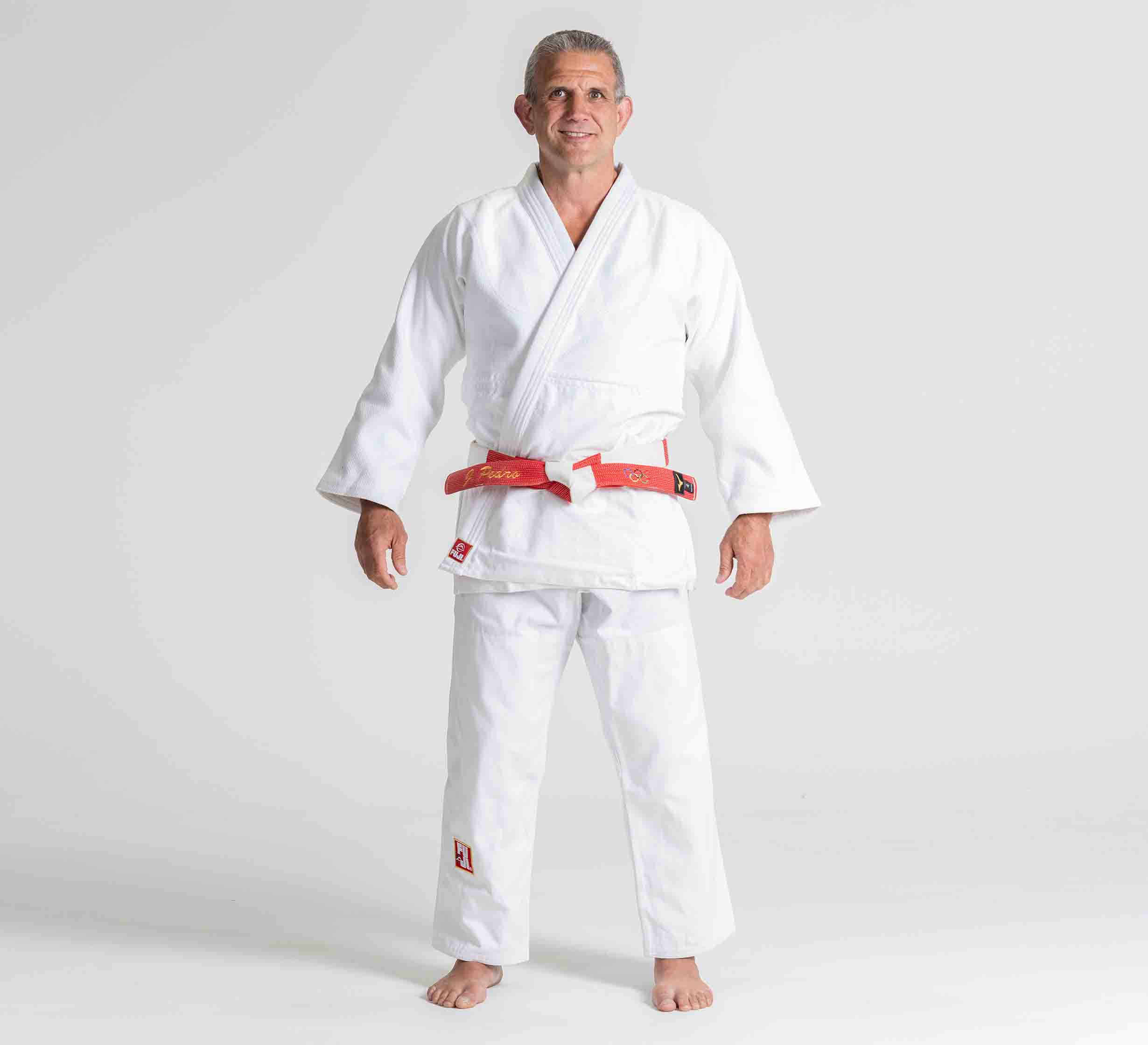 A man stands barefoot, smiling and facing forward on a white background, wearing the Euro Competition Judo Gi White with a red belt.