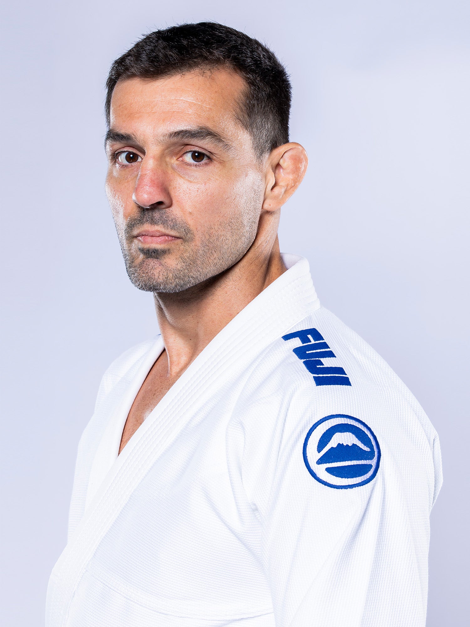 A man with short dark hair and light stubble wears the Baseline 2.0 BJJ Gi White Navy, featuring blue FUJI branding and a mountain logo on the sleeve. He looks seriously at the camera against a light background.