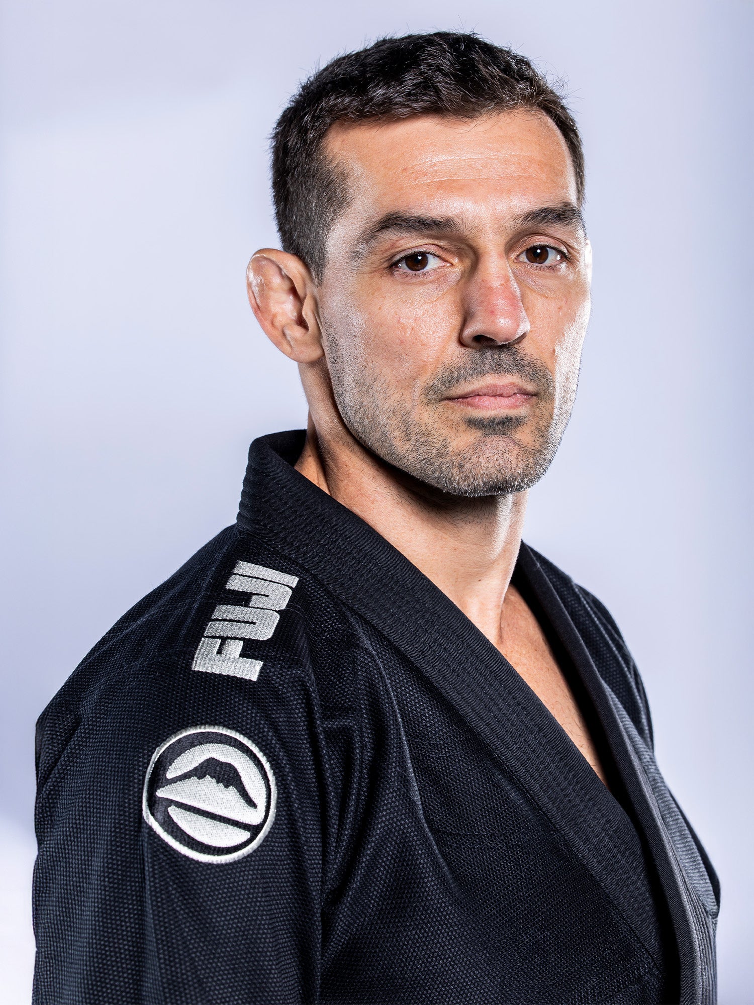 A man with short dark hair wears the Baseline 2.0 BJJ Gi Black Grey with patches, looking confidently at the camera against a plain light background.