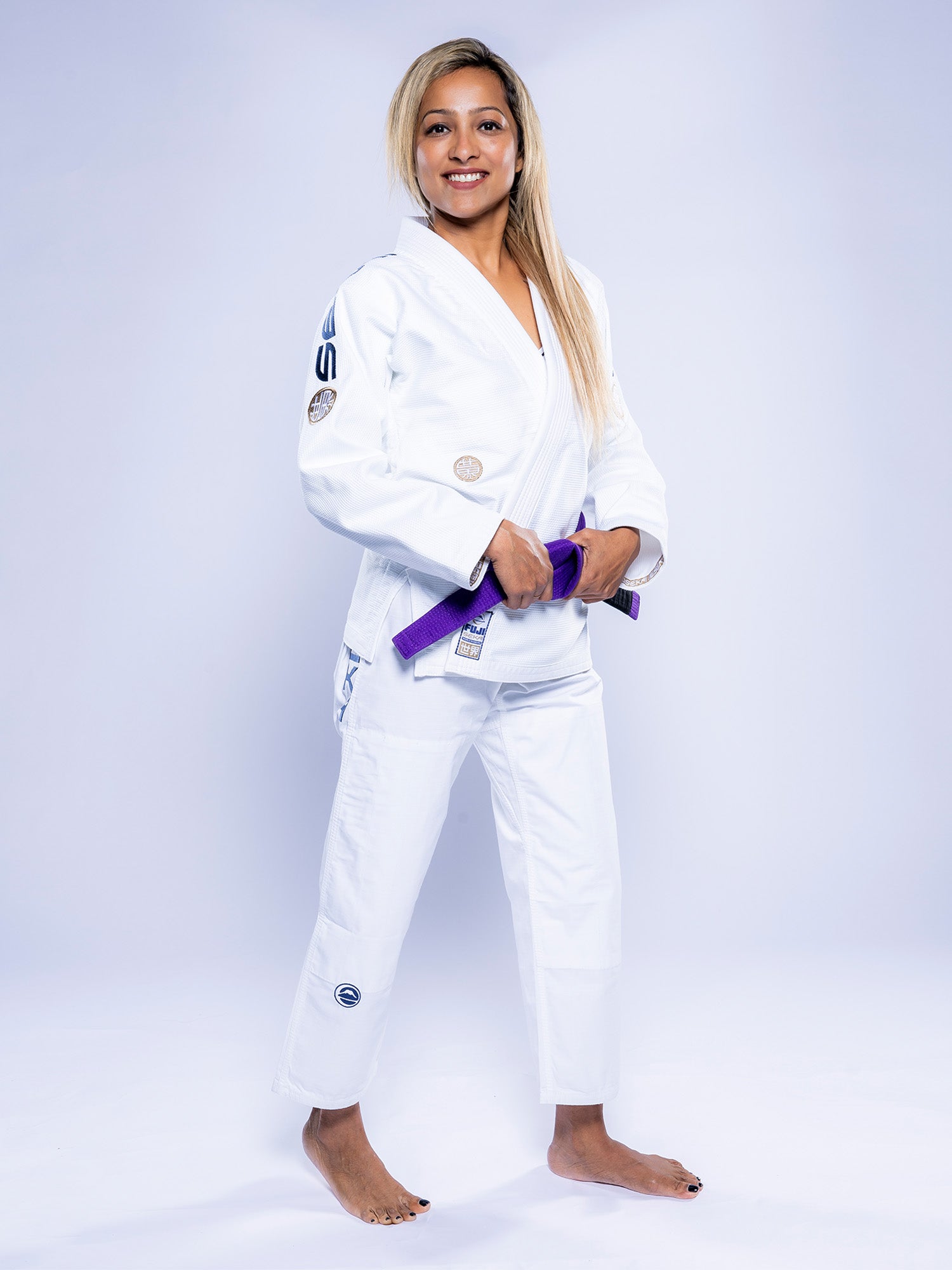 A woman wearing the Womens SEKAI 3.0 BJJ Gi White Navy stands barefoot, smiling and holding a purple belt in front of her against a plain light background.
