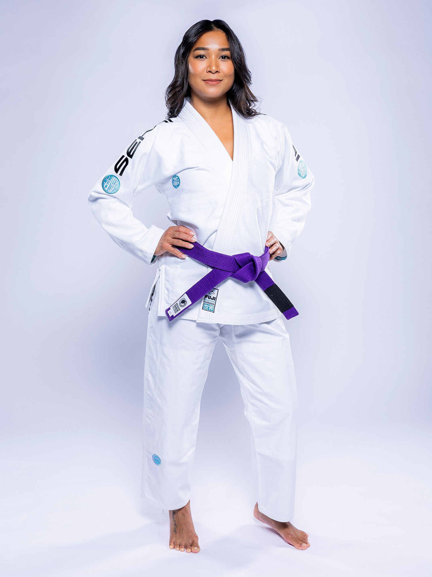 A woman stands confidently in the Womens SEKAI 3.0 BJJ Gi White Teal with a purple belt, barefoot and hands on hips, against a plain white background.