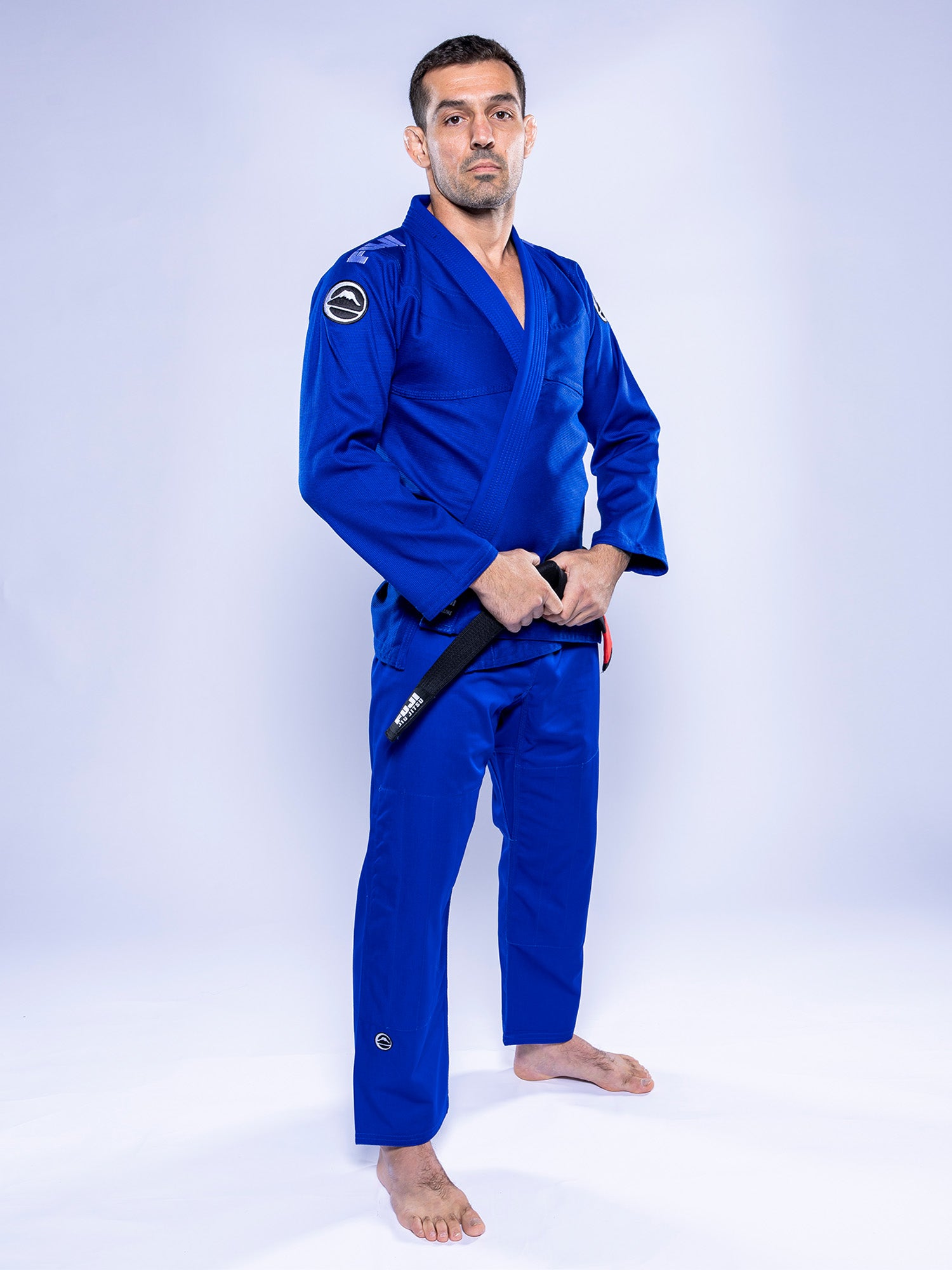 A man wears the Baseline 2.0 BJJ Gi Blue White, stands barefoot on a white background, holds a black belt in front of him, and looks at the camera.