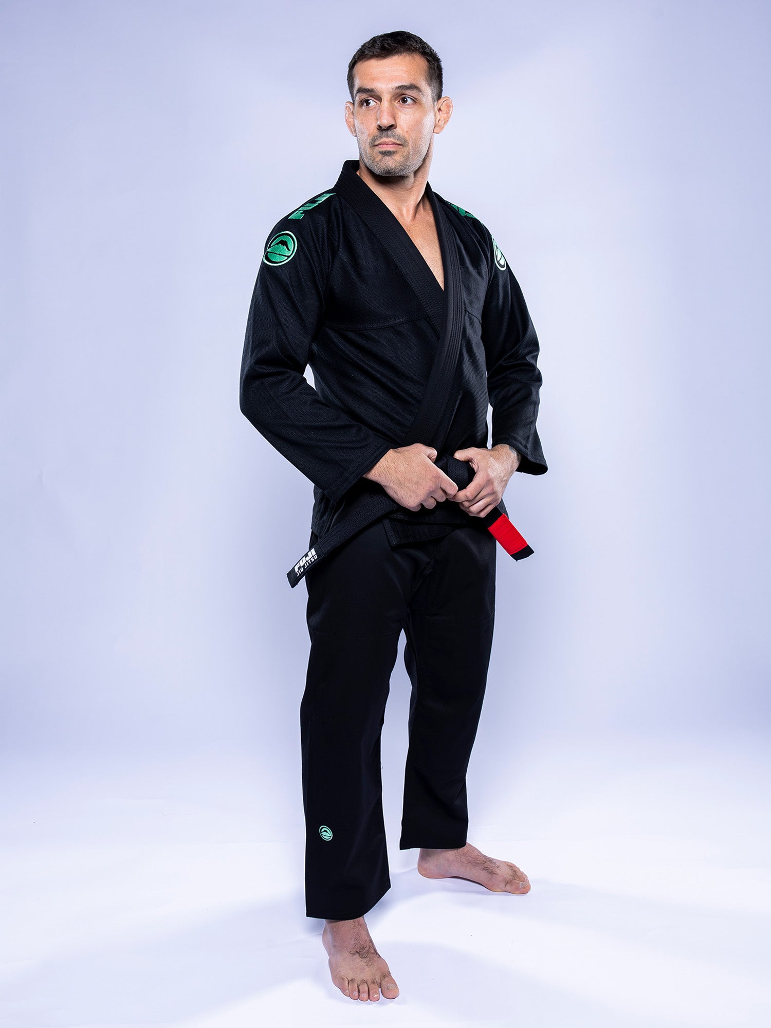 A man wears the Baseline 2.0 BJJ Gi Black Mint, barefoot on a plain light background, holding the ends of his red and black belt at his waist and looking slightly to the side with a confident expression.