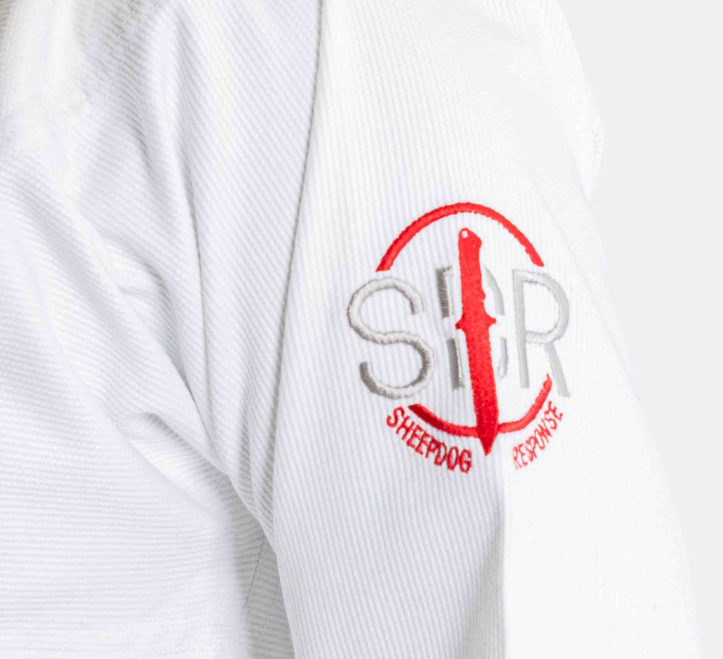 A close-up of the Suparaito BJJ Gi Sheepdog Response Edition White sleeve shows a red and gray embroidered Sheepdog Response logo with SDR letters and a knife silhouette in the center.