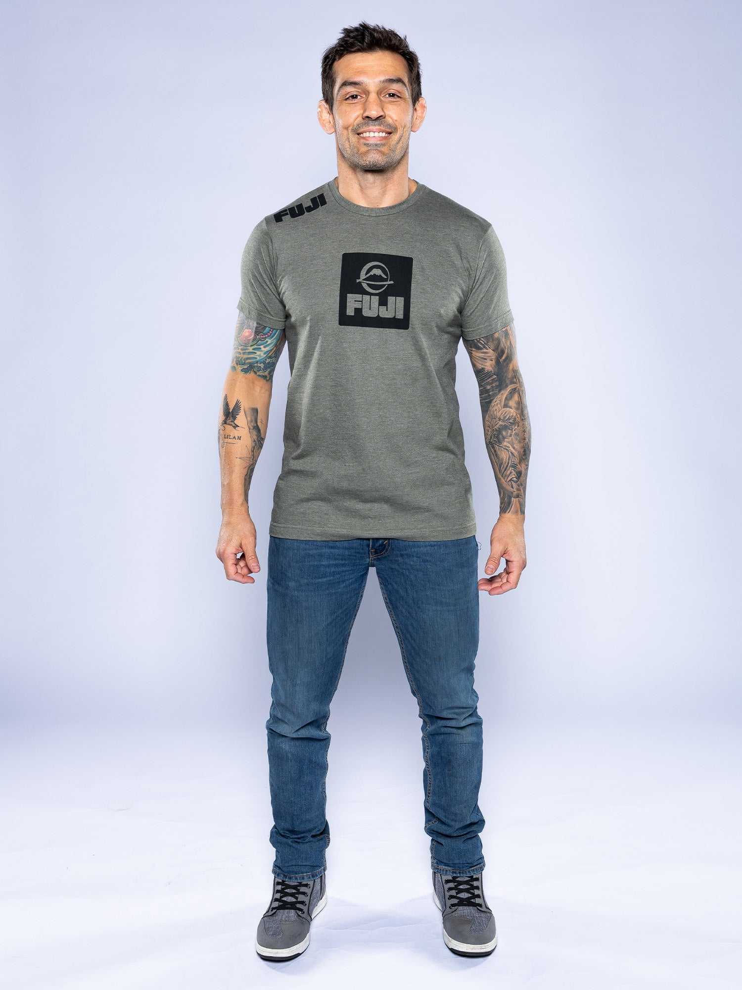 A tattooed man stands smiling in a heather grey Elevate Your Journey Shirt, blue jeans, and gray sneakers against a plain light background.