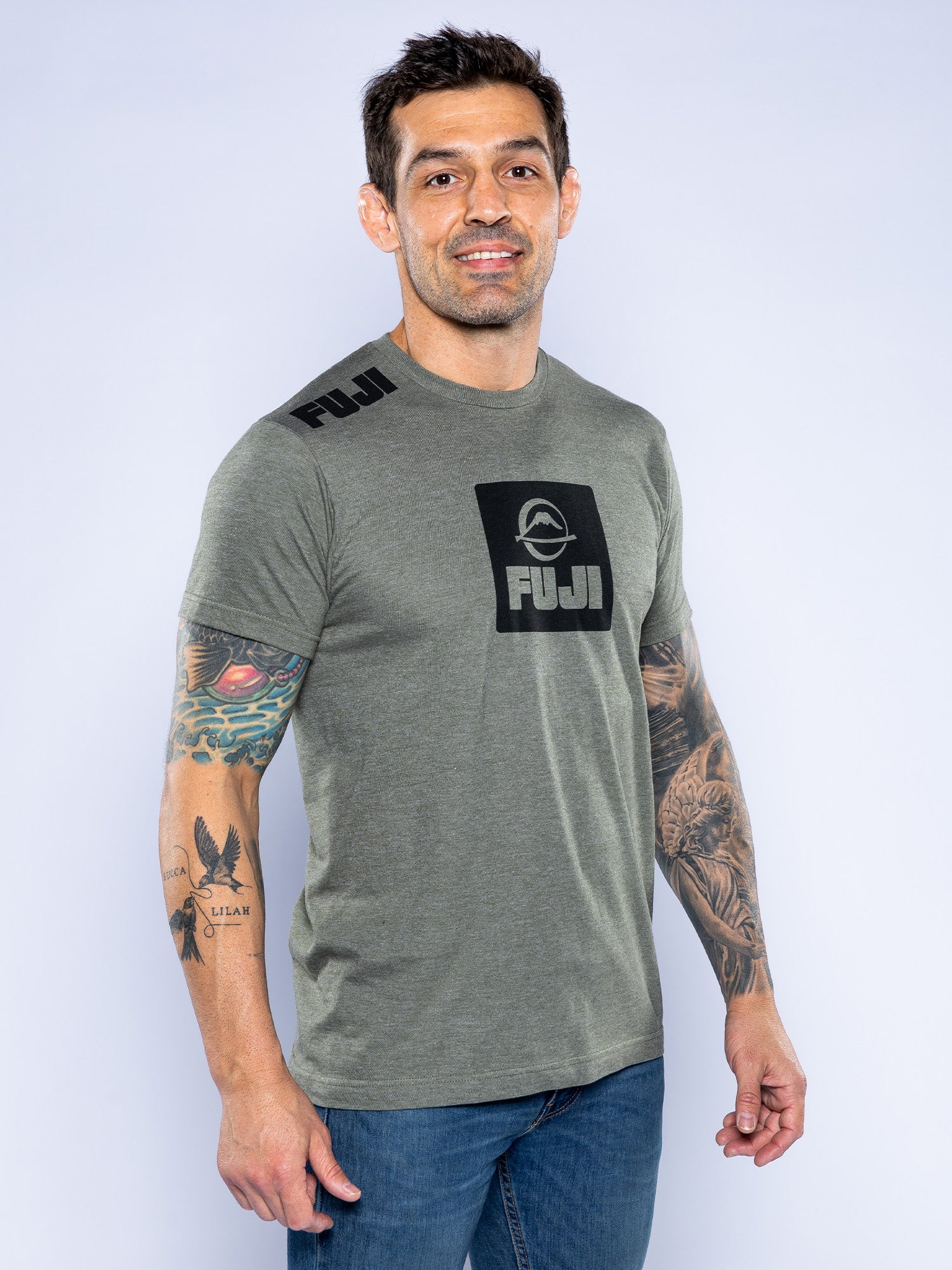 A man with short dark hair, wearing the Elevate Your Journey Shirt in Heather Grey and jeans, stands smiling with tattoos visible on both arms against a light gray background.