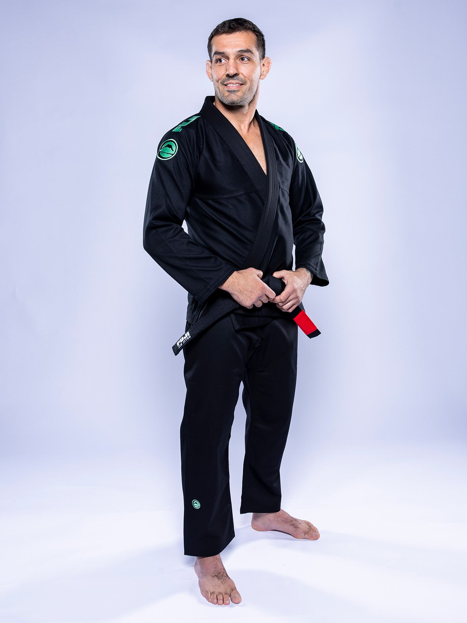 A man in the Baseline 2.0 BJJ Gi Black Mint stands barefoot on a light background, holding his belt and smiling while looking to the side.