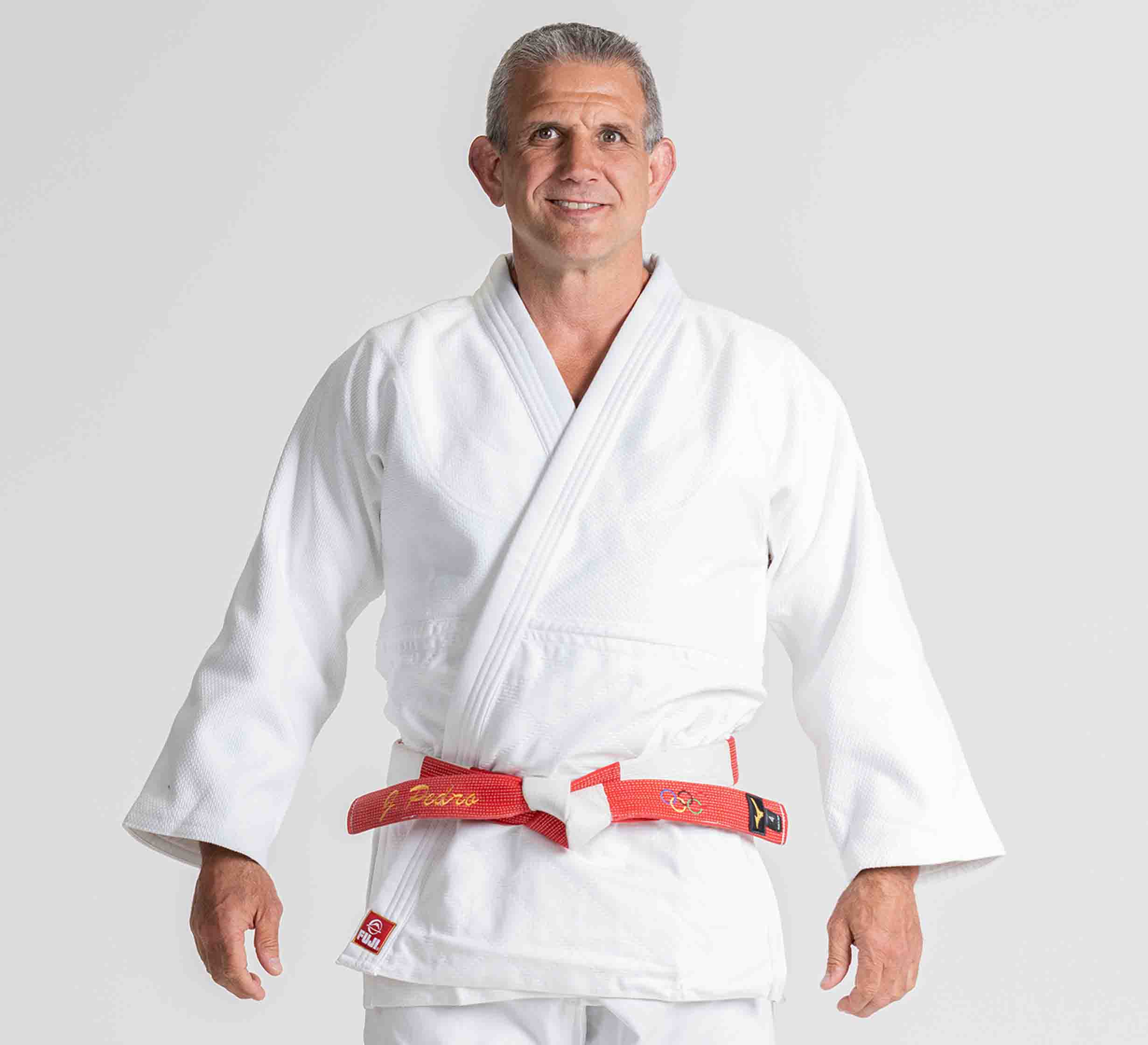 A man wearing the Euro Competition Judo Gi White and a red belt stands smiling and facing forward against a plain white background.