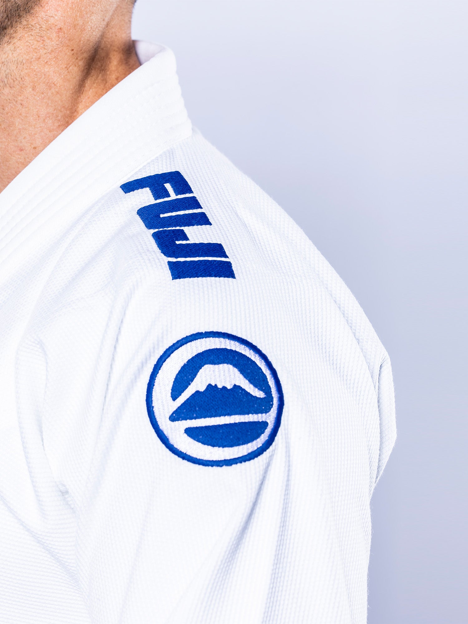 Close-up of a person wearing the Baseline 2.0 BJJ Gi White Navy, featuring blue FUJI text and a mountain logo on the shoulder. Only the upper shoulder, part of the neck, and face are visible against a light background.