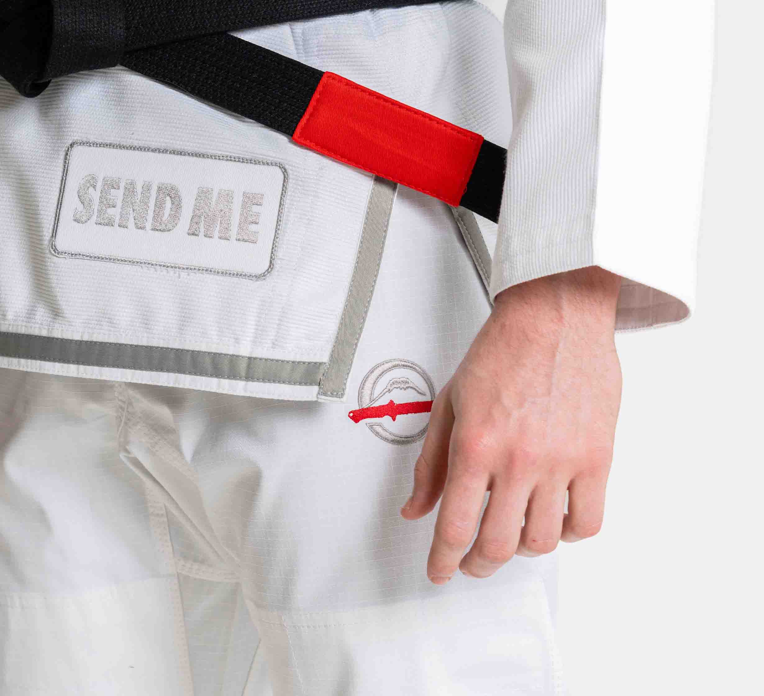 A person wearing a Suparaito BJJ Gi Sheepdog Response Edition White with "SEND ME" on the jacket and a black belt with a red tag, standing with their right hand relaxed by their side.