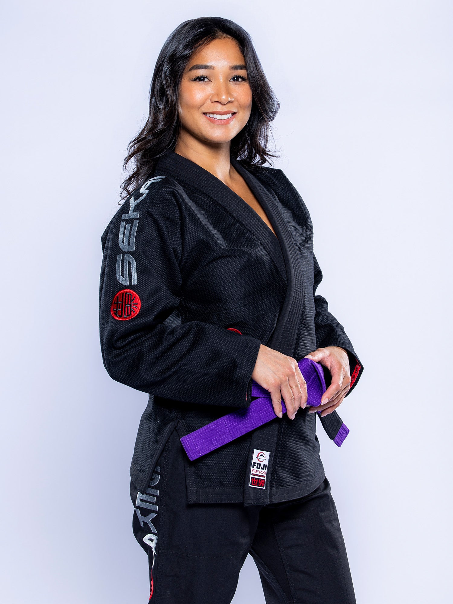 A woman in the Womens SEKAI 3.0 BJJ Gi Black Grey with patches and a purple belt smiles against a plain white background.