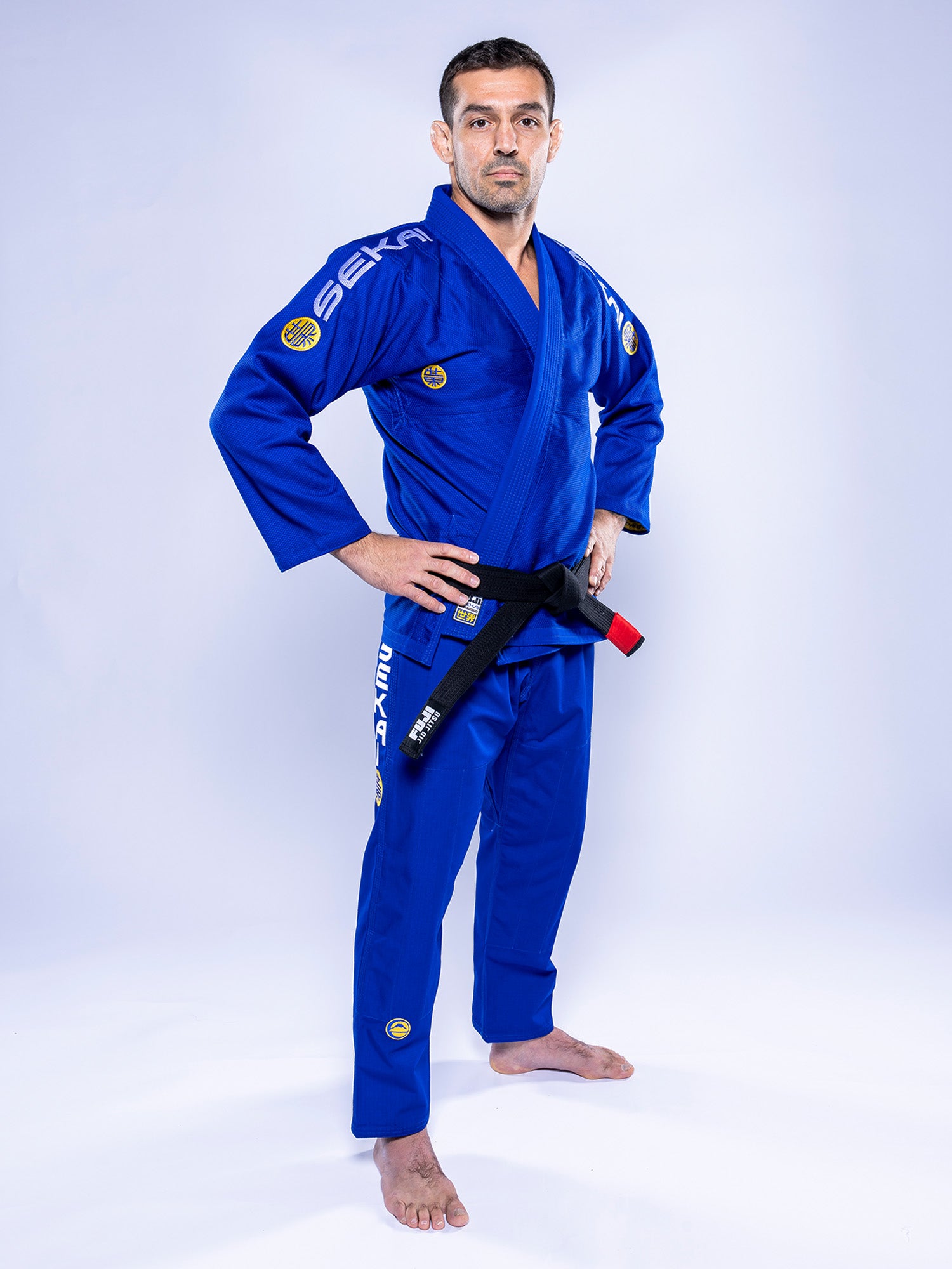 A man in a SEKAI 3.0 BJJ Gi Blue Yellow with a black belt stands barefoot on a white background, facing the camera with his hands on his hips.