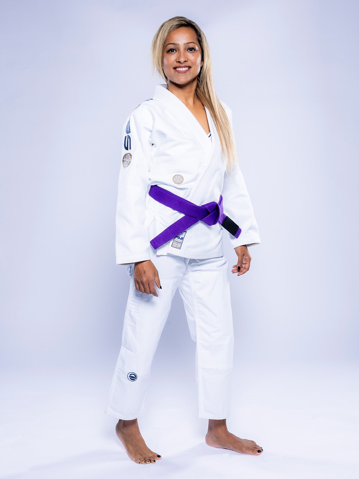 A woman with long loose hair smiles confidently at the camera, barefoot on a light background, wearing the white Women's SEKAI 3.0 BJJ Gi in Navy with a purple belt.
