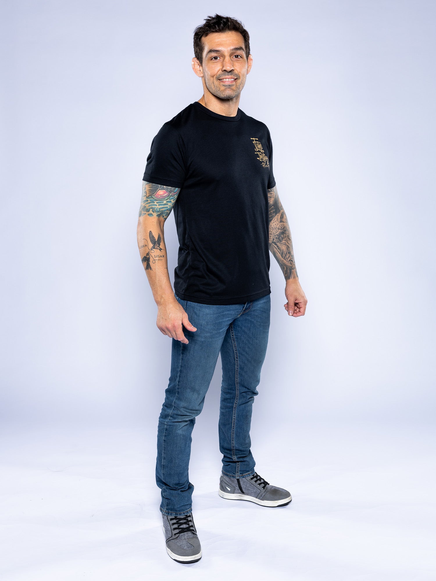 A man with short dark hair and tattoos on both arms stands smiling, wearing the Japanese Street Art Shirt Black, blue jeans, and gray sneakers against a plain light background.