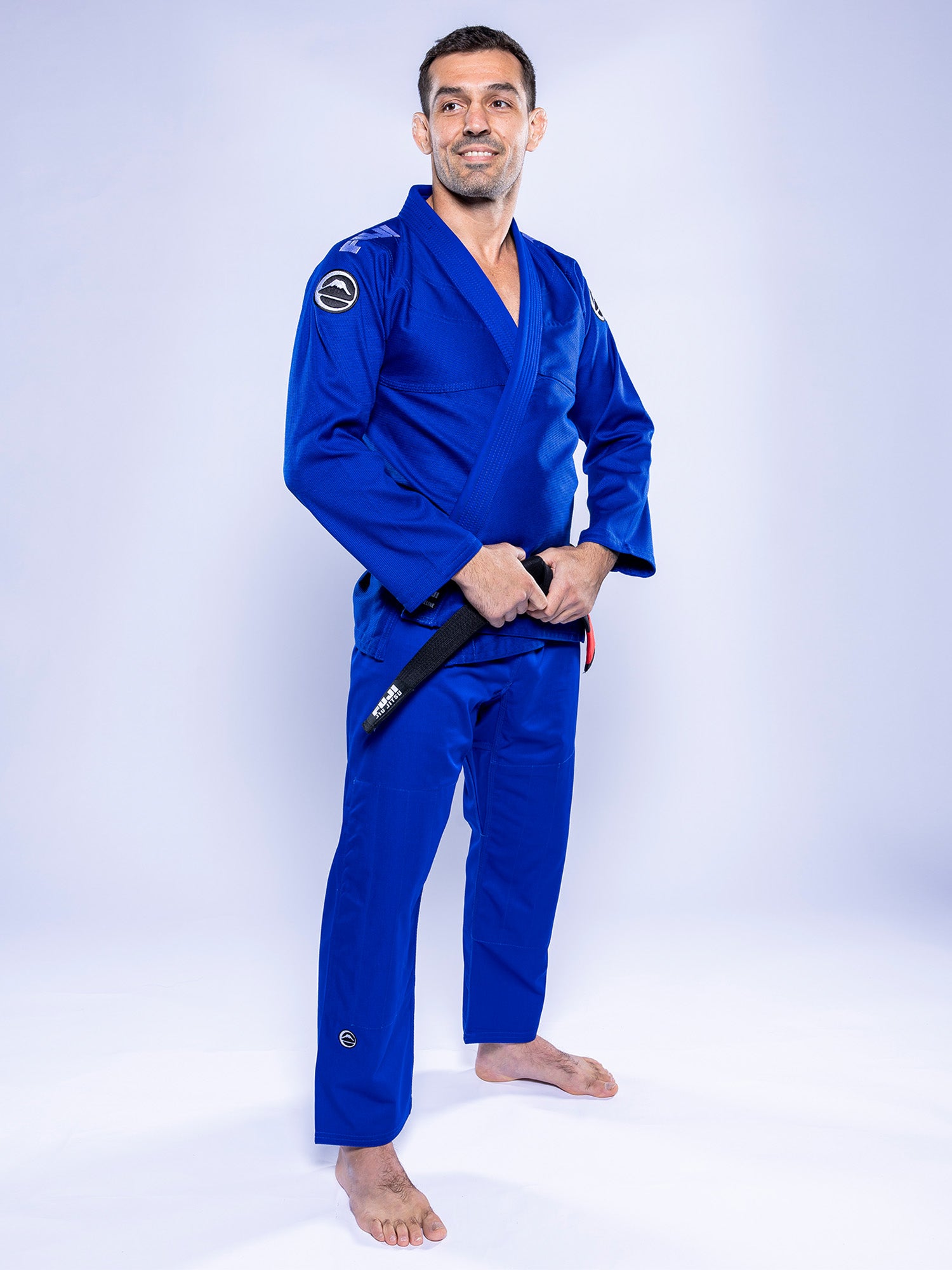 A man in the Baseline 2.0 BJJ Gi Blue White stands barefoot on a white background, holding a black belt and smiling slightly while looking off to the side.