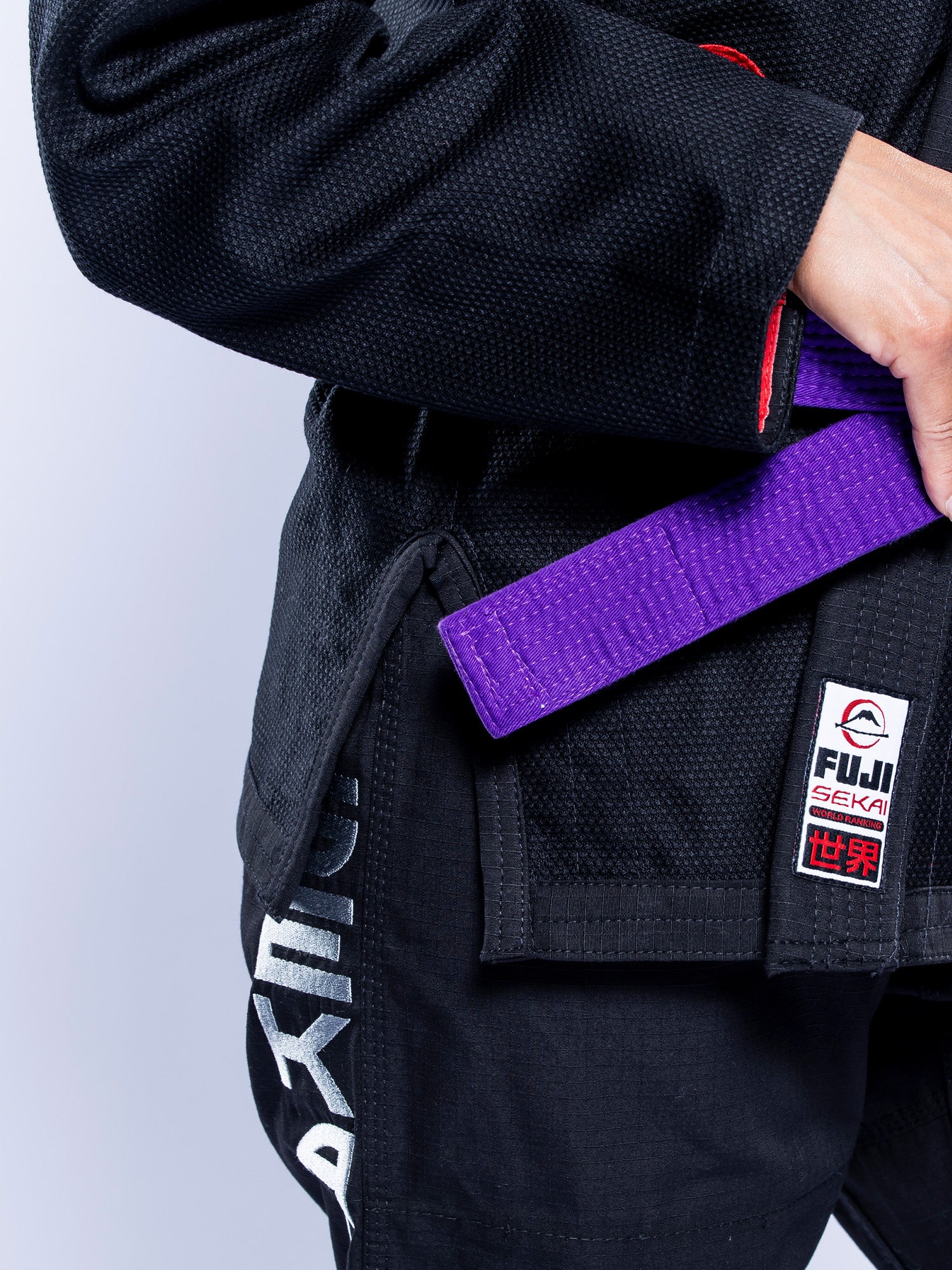 A woman wearing the Womens SEKAI 3.0 BJJ Gi Black Grey with white lettering on the pants and a purple belt adjusts her belt. The Fuji brand patch is visible on the gi.