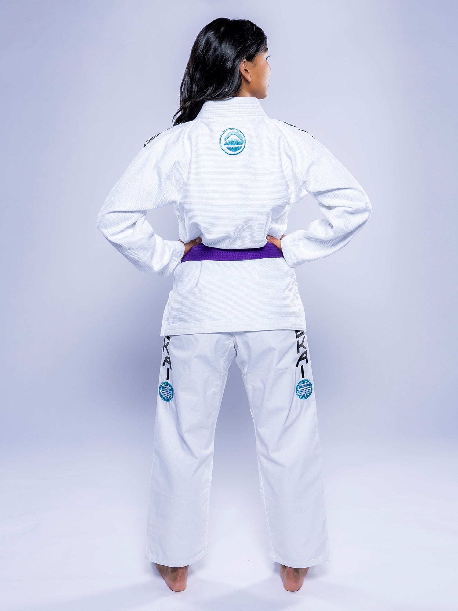 A woman with long dark hair stands barefoot, facing away, wearing the Womens SEKAI 3.0 BJJ Gi in white with teal accents and a purple belt. Blue patches and black lettering are on the pants. The background features a light gradient.