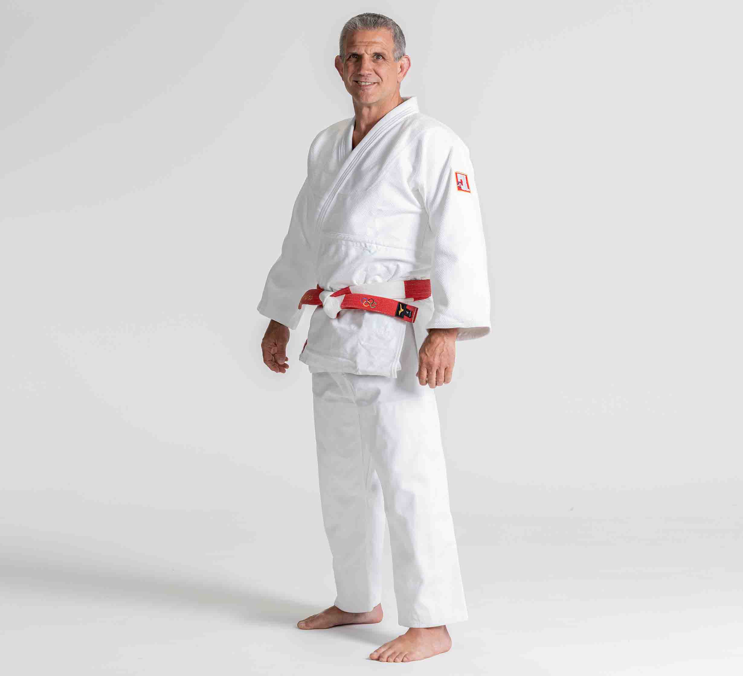 A man stands barefoot on a plain background, wearing the Euro Competition Judo Gi White with a red and black belt, smiling slightly and facing the camera.