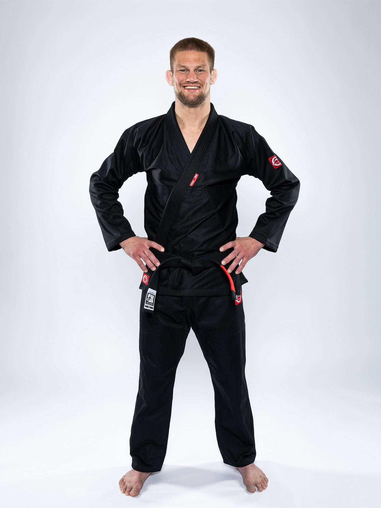 A man wearing the Ultra Lite 300 BJJ Gi Black stands barefoot on a white background, smiling with his hands on his hips.