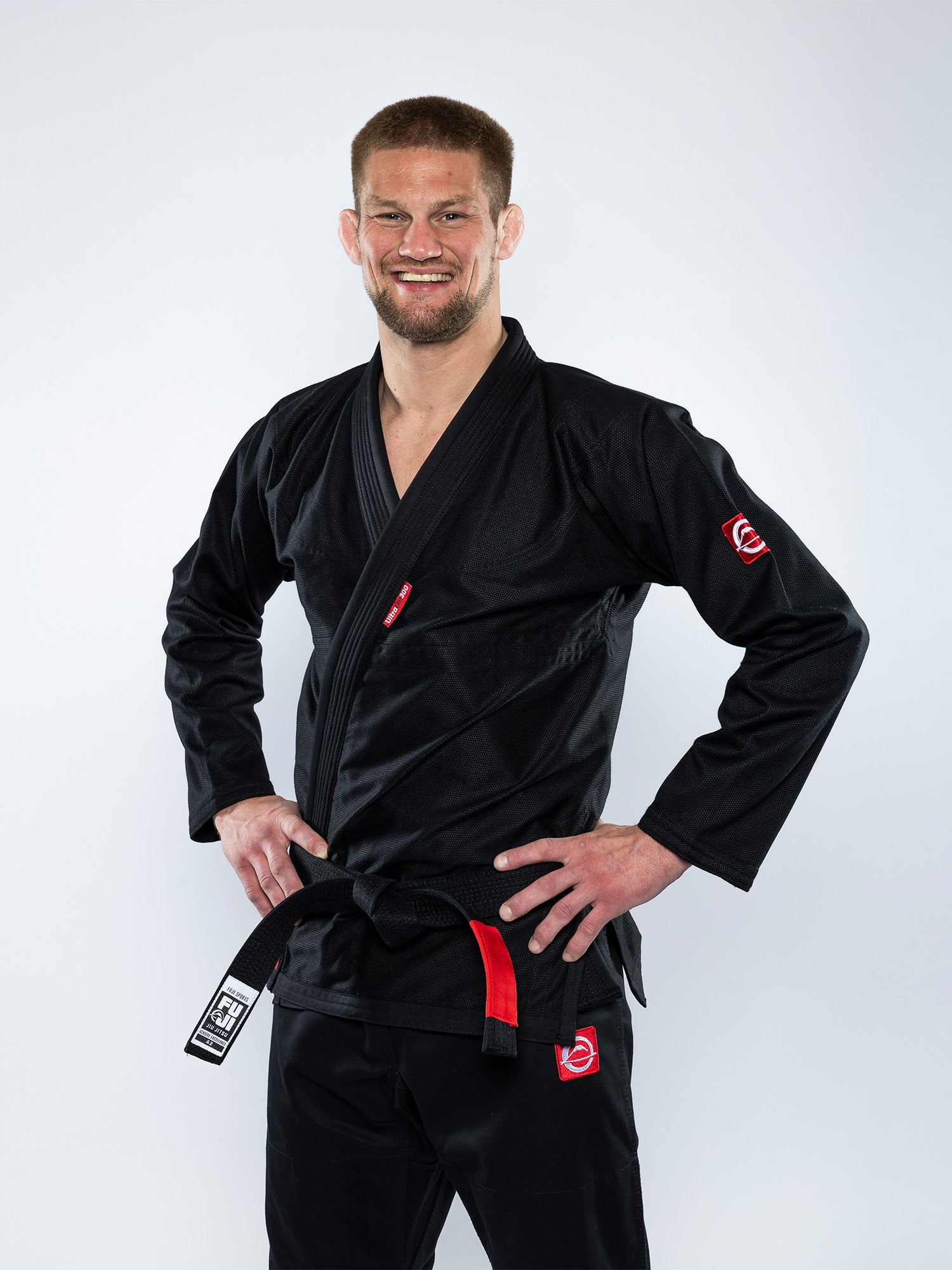A man wears the Ultra Lite 300 BJJ Gi Black with a black belt, smiling with his hands on his hips against a plain light background.