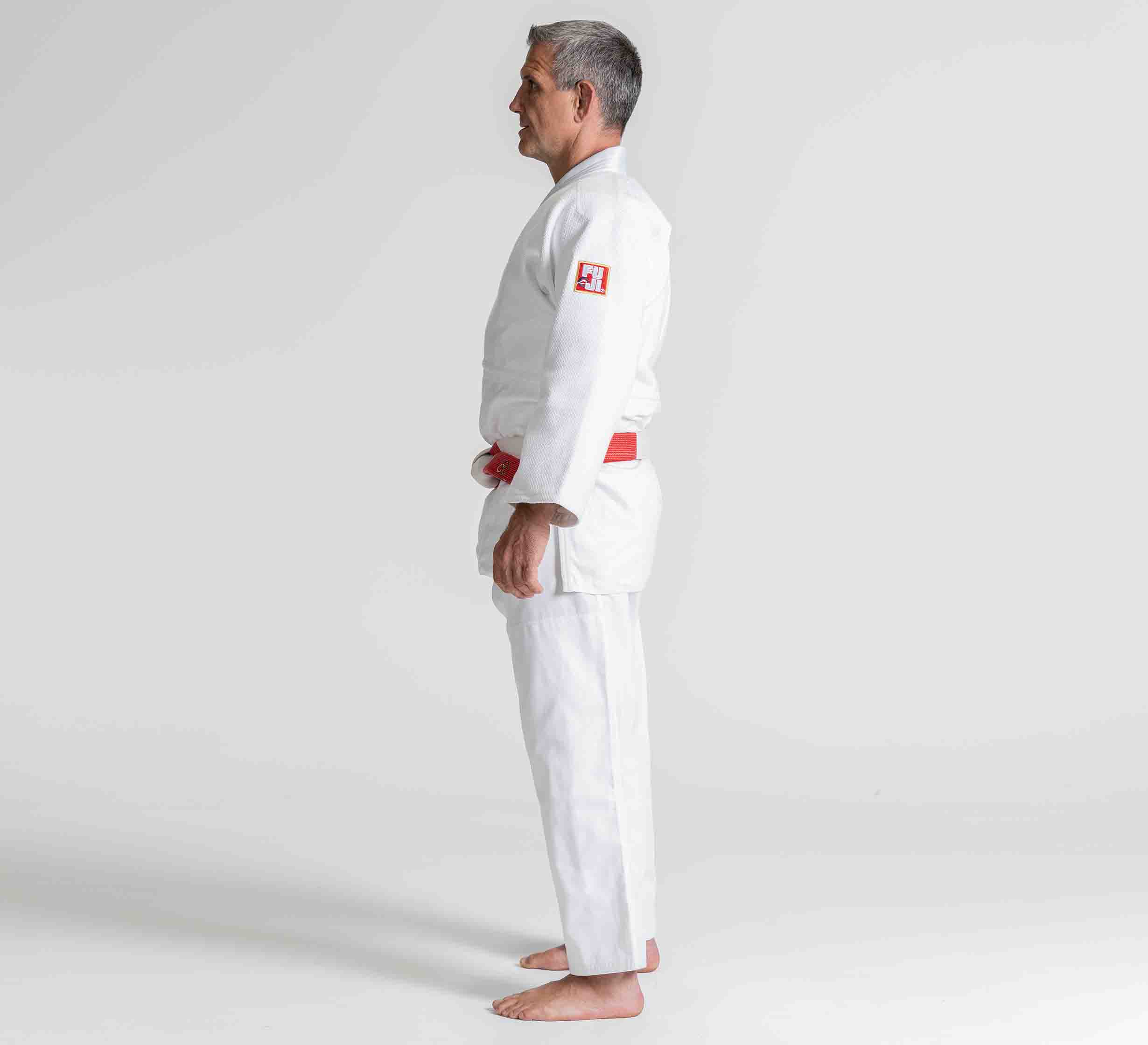 A man stands barefoot in profile against a plain white background, wearing the Euro Competition Judo Gi White with a red and white belt.