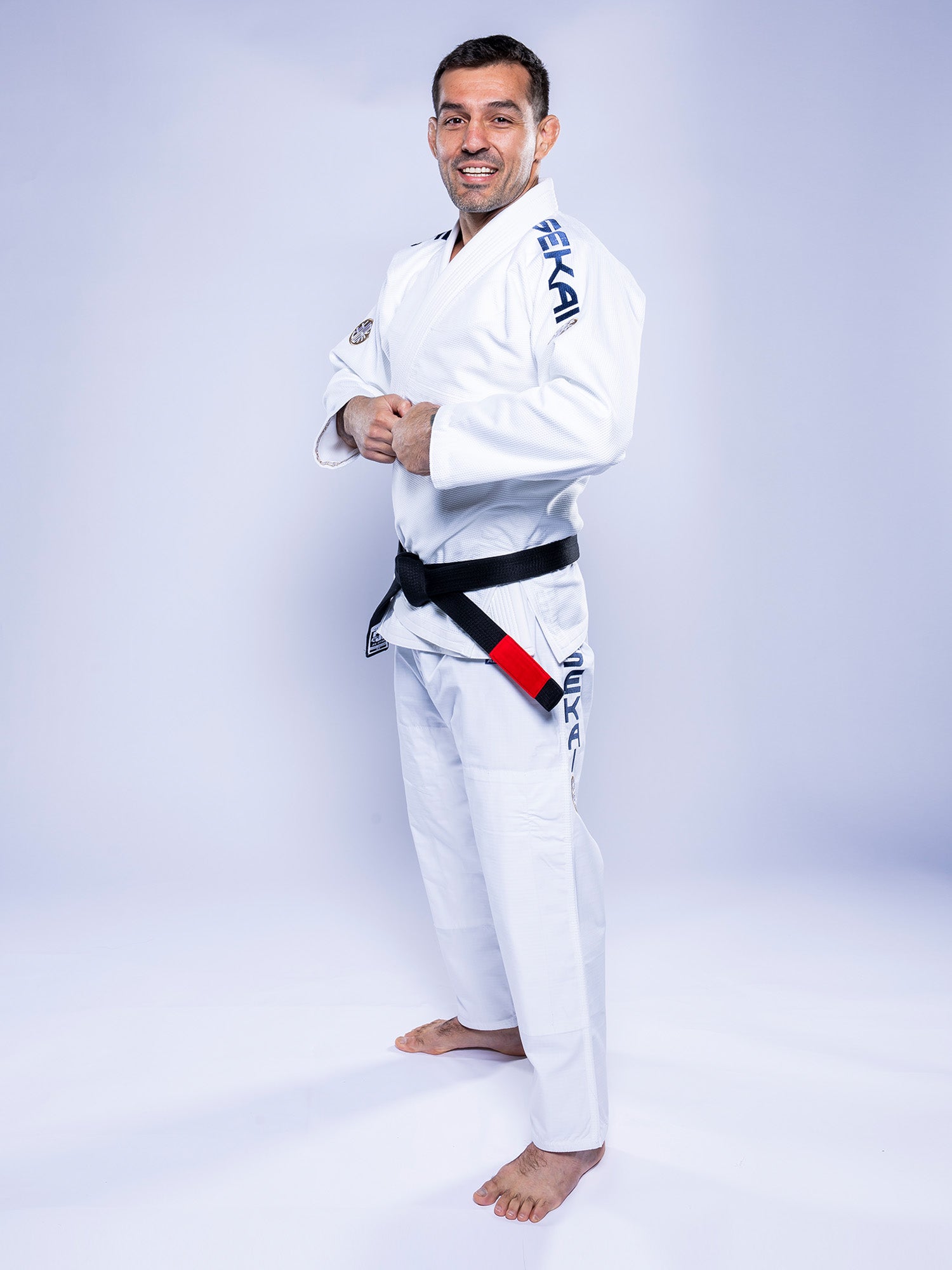 A man stands barefoot on a light background, smiling with his hands clasped at his waist, wearing the SEKAI 3.0 BJJ Gi White Navy and a black belt.