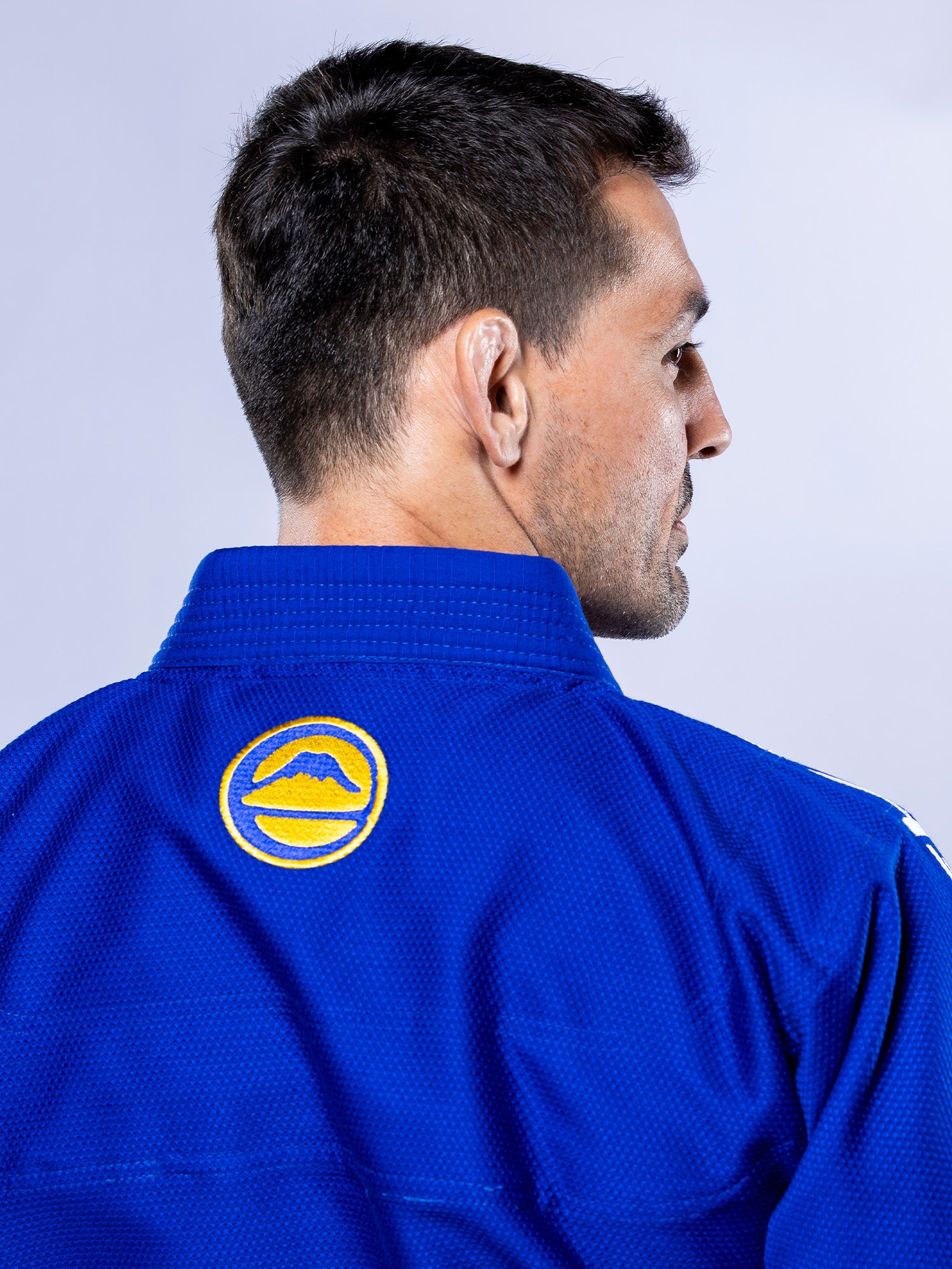 A man with short dark hair is seen from behind, wearing the SEKAI 3.0 BJJ Gi Blue Yellow, which features a yellow and blue circular patch on the back collar. The background is light gray.