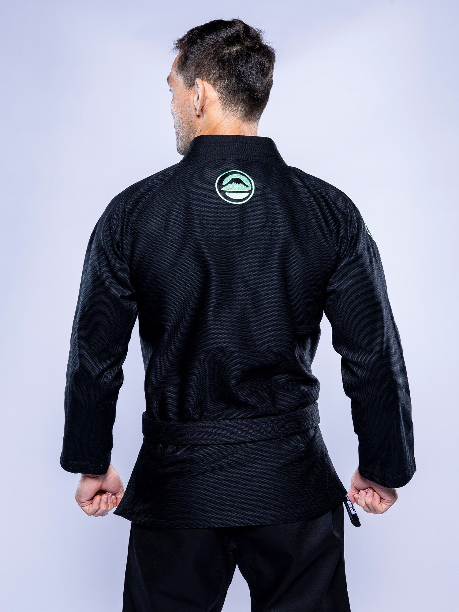 A person stands with their back to the camera, wearing the Baseline 2.0 BJJ Gi Black Mint, featuring a green circular logo on the upper back and a black belt tied at the waist.