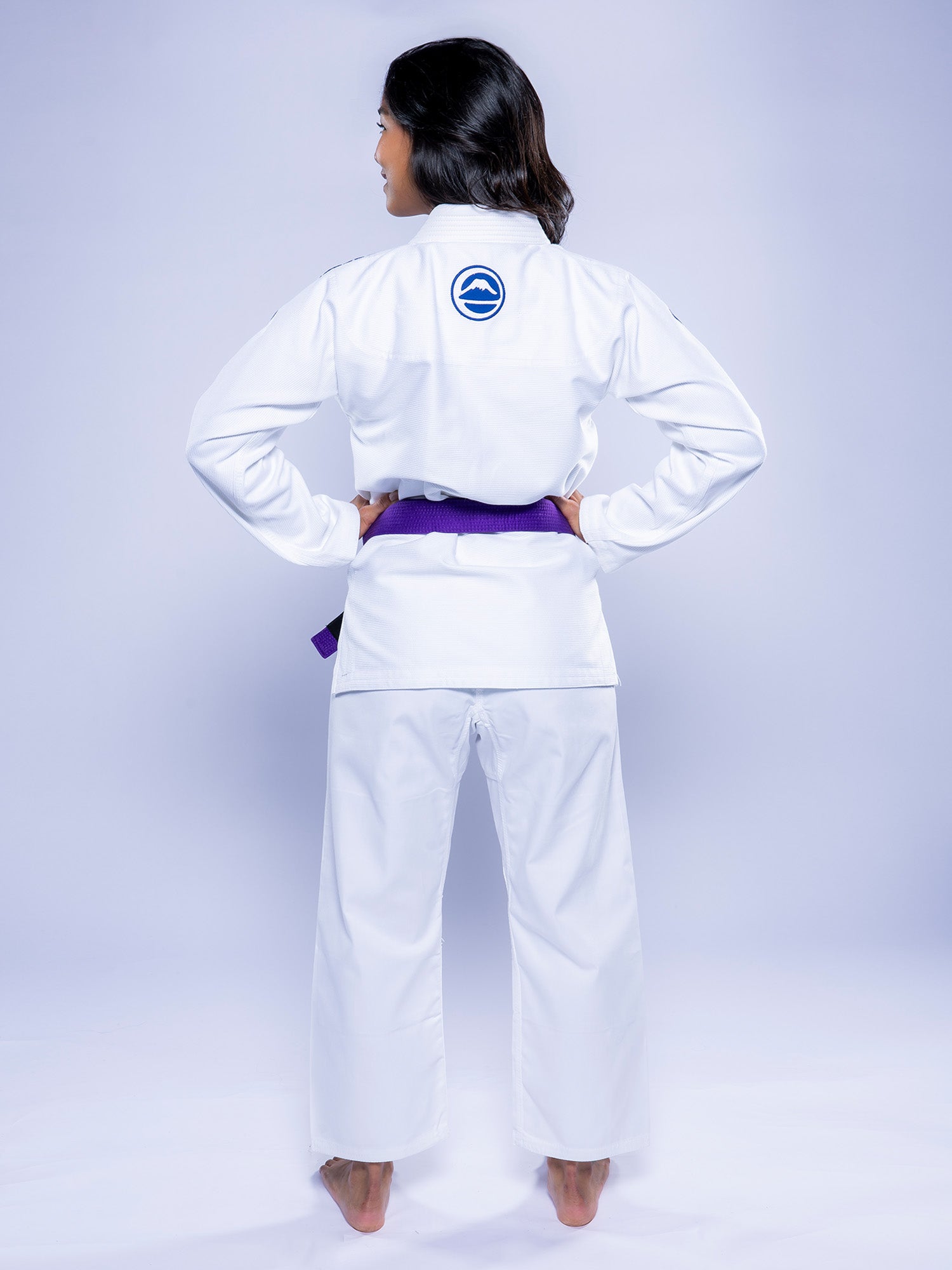 A woman in the Womens Baseline 2.0 BJJ Gi White Navy, featuring a circular blue back logo, stands barefoot facing away with hands on hips against a plain light background. She wears a purple belt.