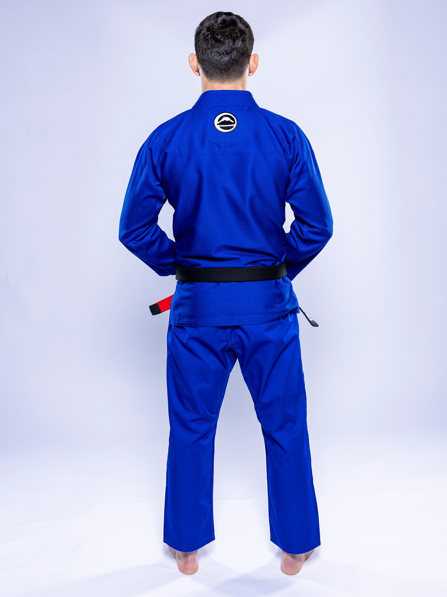 A person stands barefoot on a white surface, facing away, wearing the Baseline 2.0 BJJ Gi Blue White with a black belt and a circular patch on the upper back against a plain light-colored background.