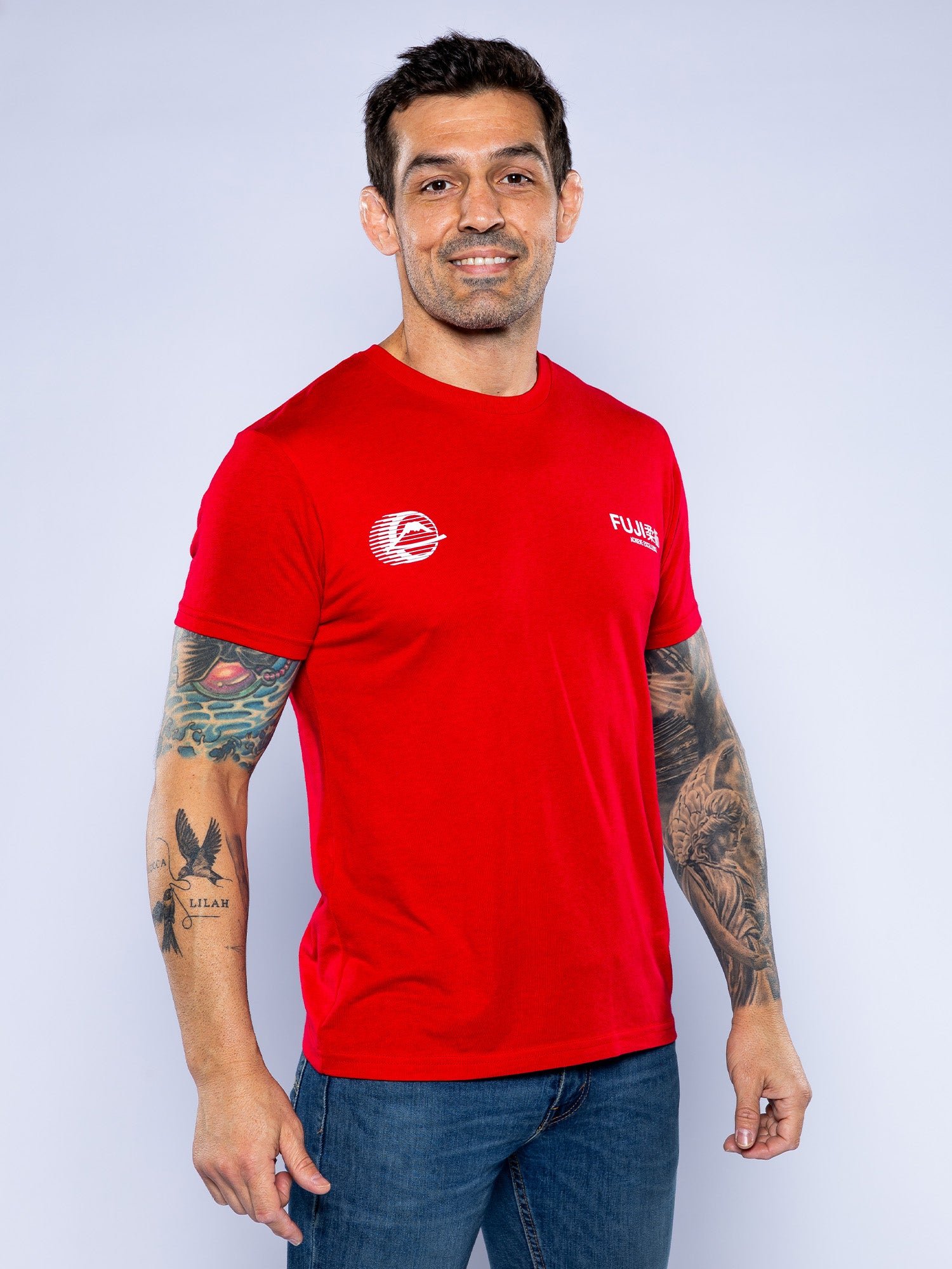 A man with short dark hair, tattoos on both arms, and a beard smiles while wearing the Iconic Jiu Jitsu Shirt Red and blue jeans, standing against a plain light background.