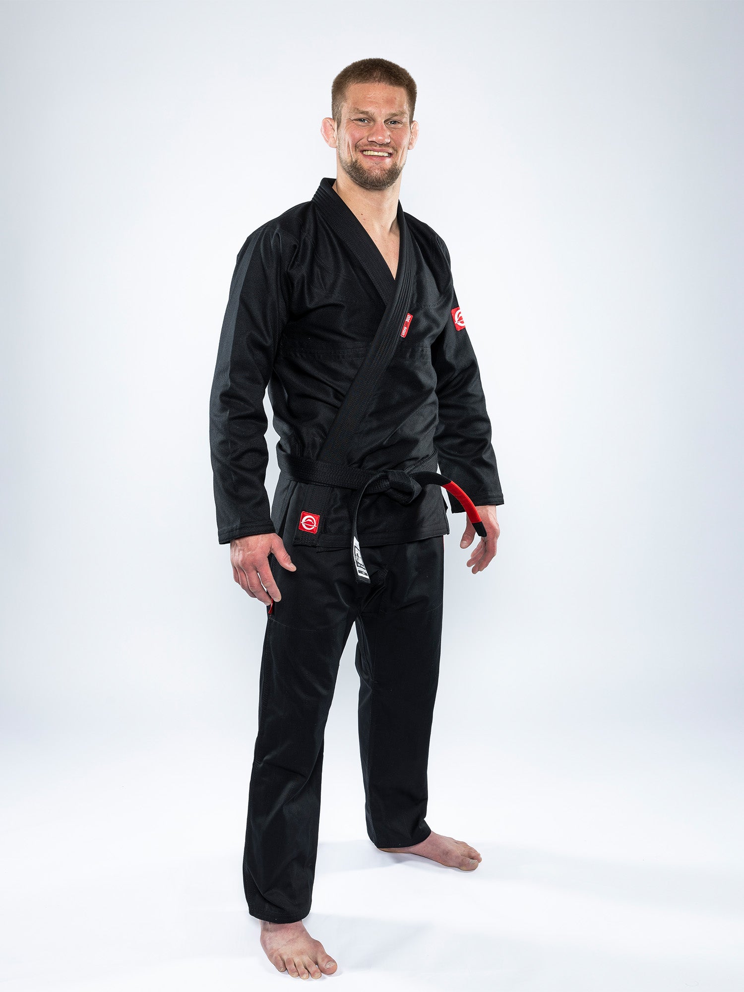 A barefoot, smiling man with short hair and a beard stands against a plain white background wearing the Ultra Lite 300 BJJ Gi Black with a black belt.