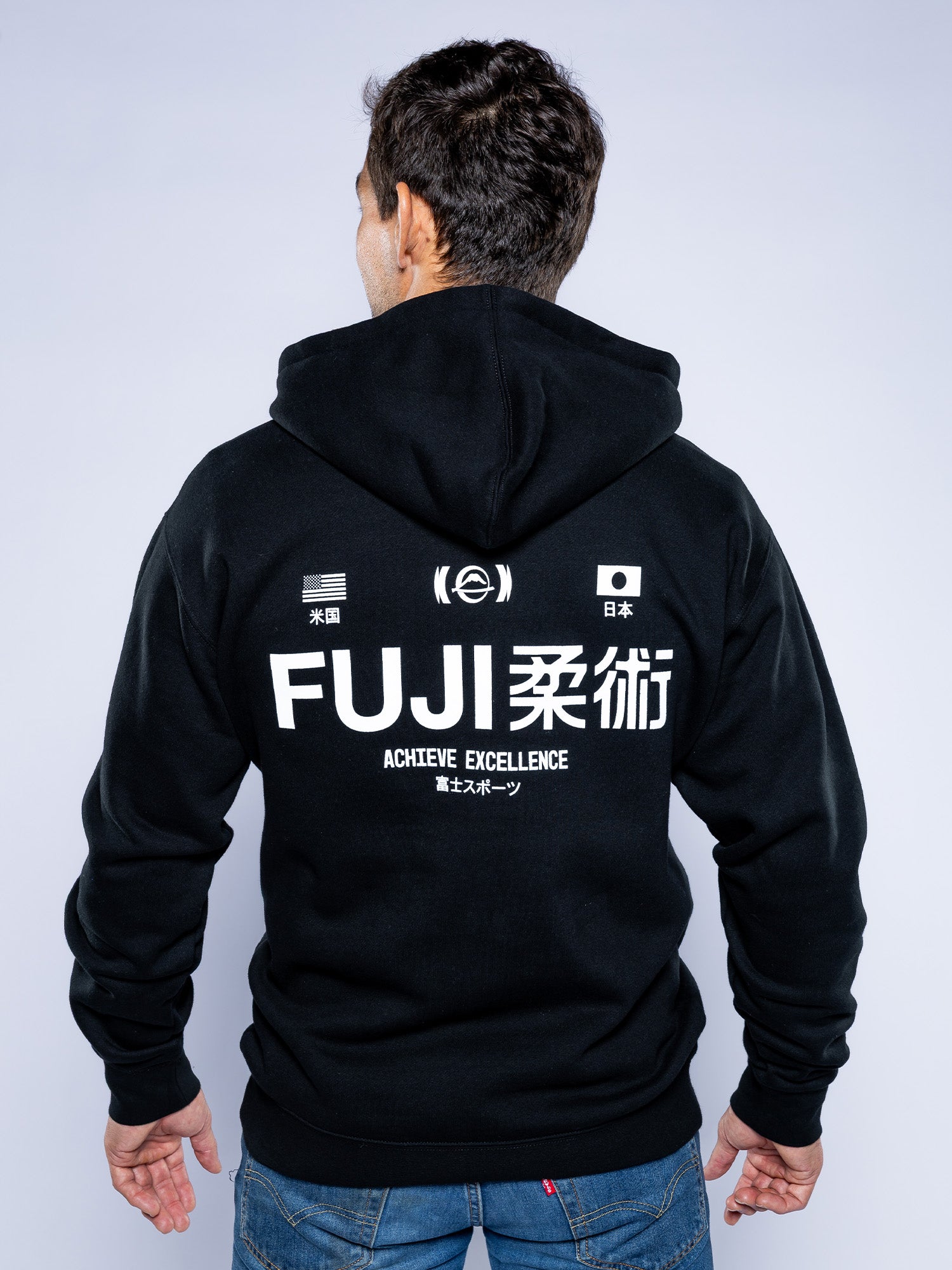 A person faces away, wearing the Iconic Jiu Jitsu Hoodie Black featuring white text "FUJI 柔術 ACHIEVE EXCELLENCE," small US and Japan flags, and additional Japanese characters.