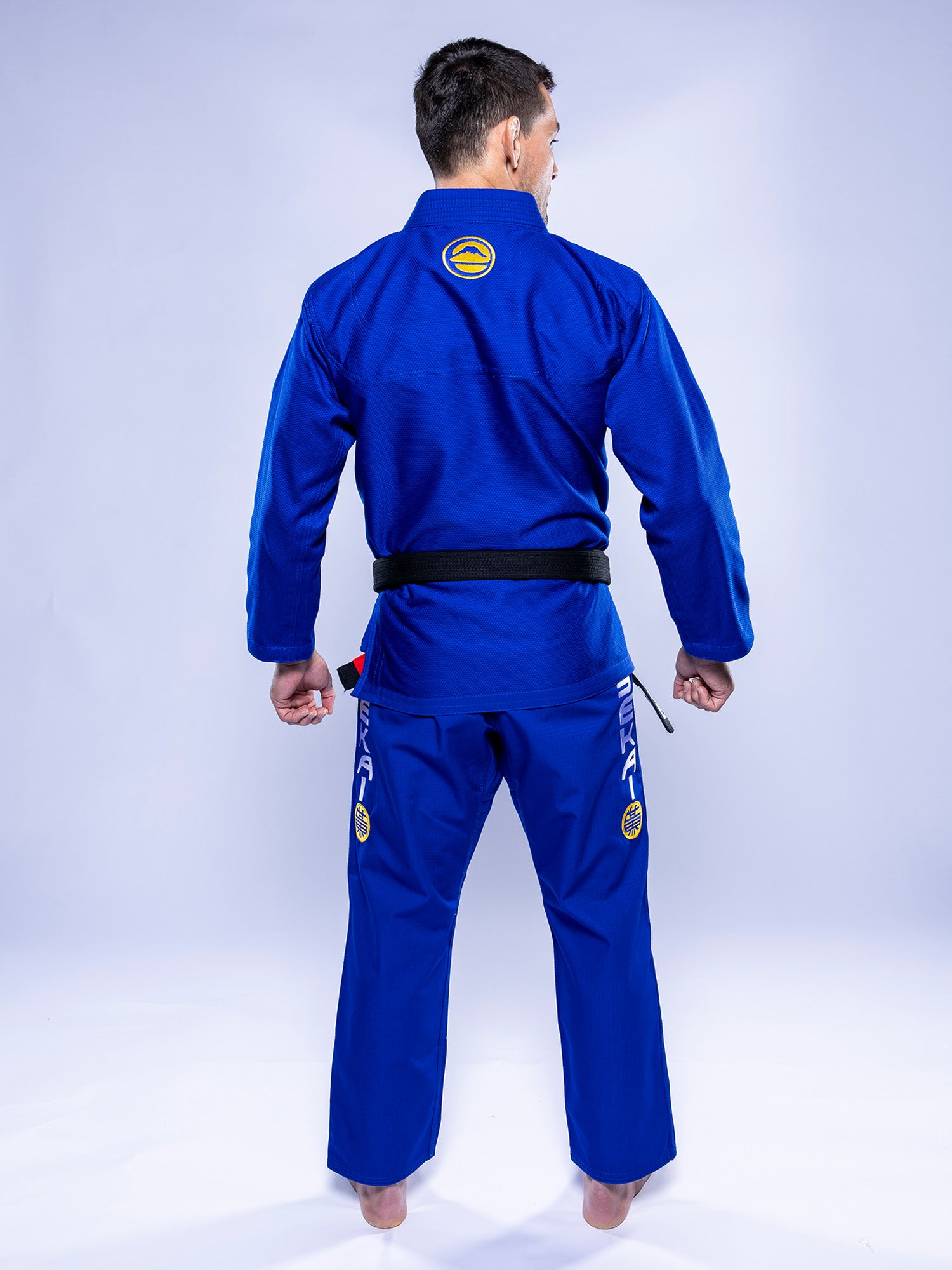 A person wearing the SEKAI 3.0 BJJ Gi Blue Yellow with a black belt stands barefoot, facing away to display the back of the uniform, which features patches and logos on both the jacket and pants.