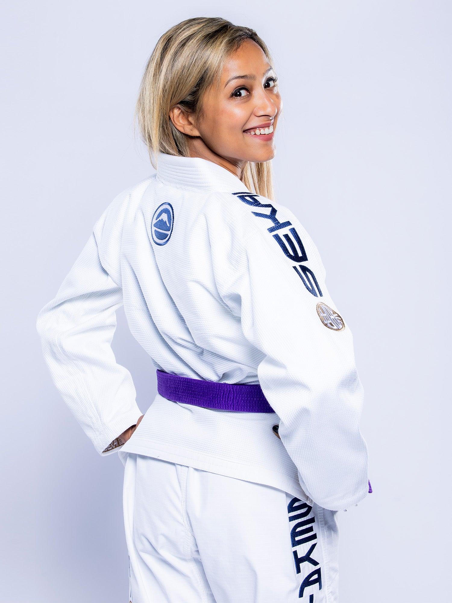 A woman with long blonde hair smiles over her shoulder, wearing the Womens SEKAI 3.0 BJJ Gi White Navy with a purple belt and patches on the sleeves and back, standing against a light background.