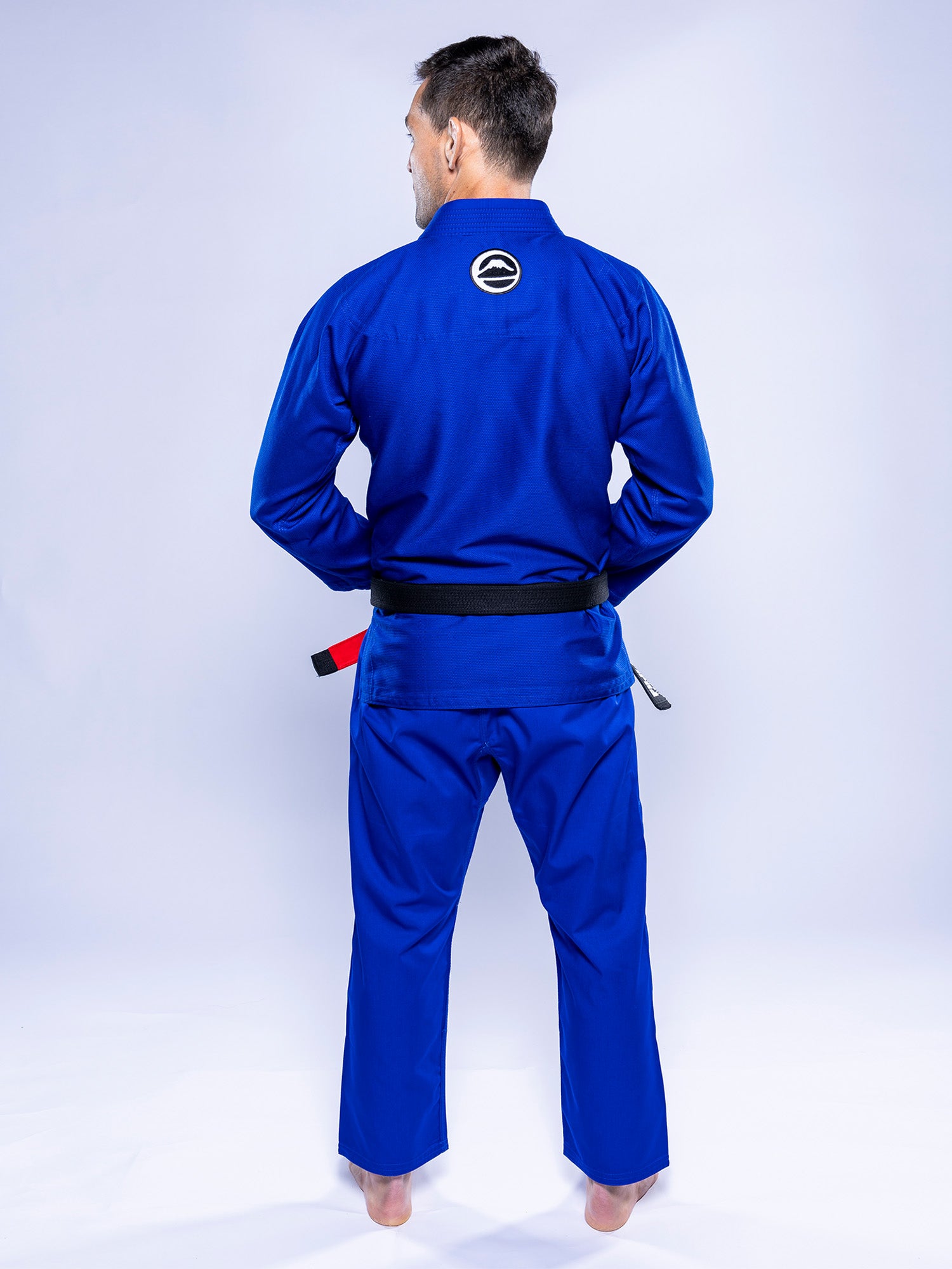 A man stands barefoot, facing away from the camera, in a Baseline 2.0 BJJ Gi Blue White with a black belt and a circular patch on the upper back of his uniform.