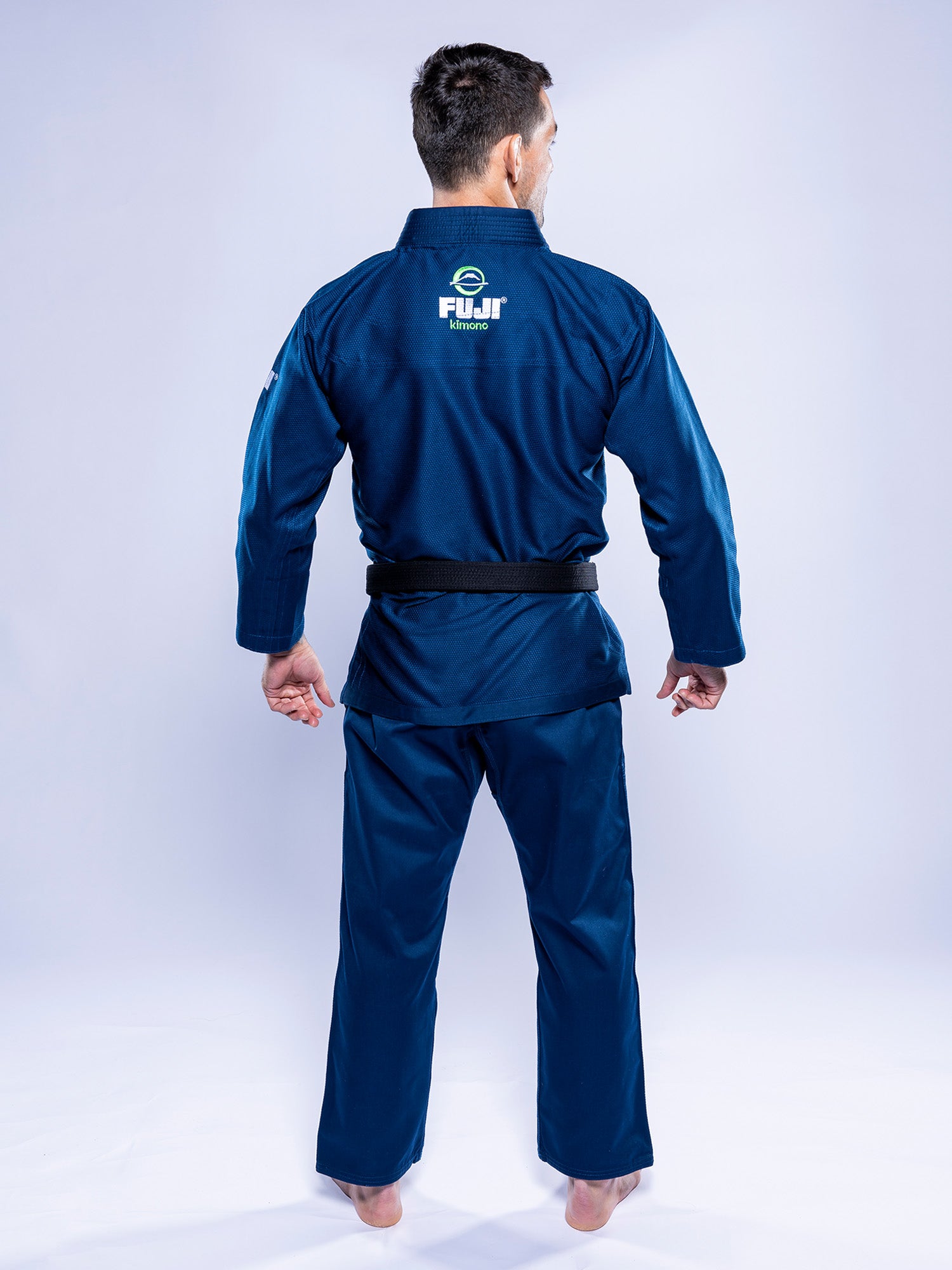 A person stands barefoot, facing away, wearing the Lightweight BJJ Gi Navy with a black belt. “FUJI Kimonos” is written on the back. The background is plain and light-colored.