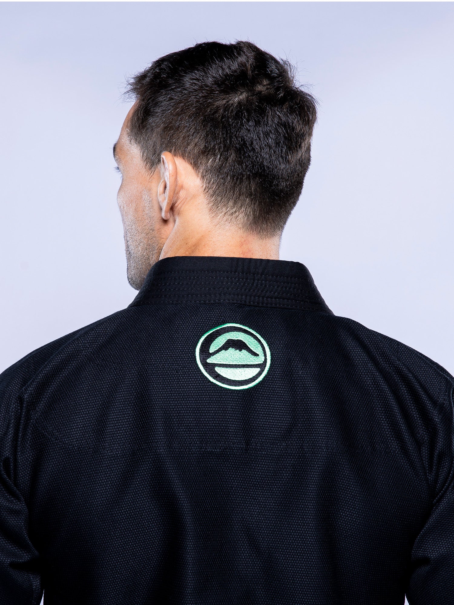 A man seen from behind wears the Baseline 2.0 BJJ Gi Black Mint, featuring a green circular mountain logo on the back of the collar. The background is plain and light-colored.