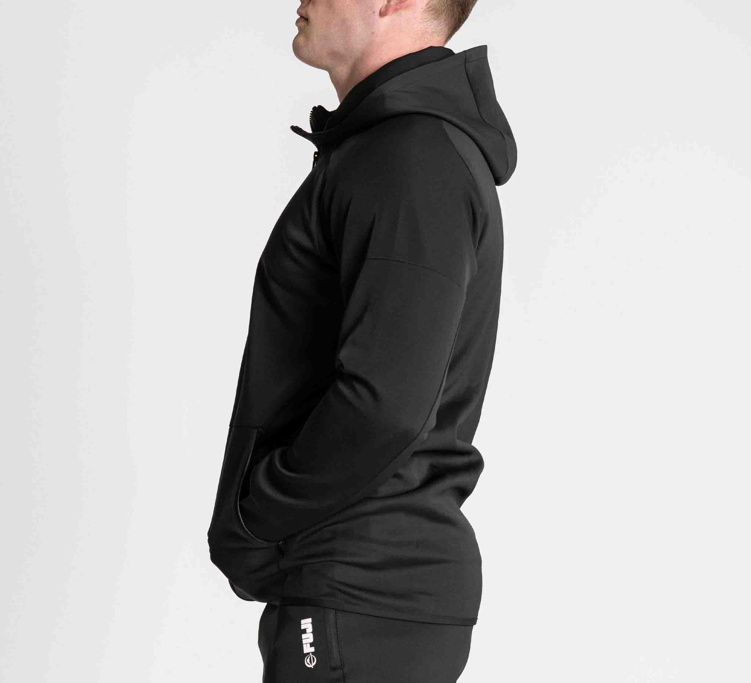 A person stands in profile against a white background wearing the FUJI Performance Jacket Black and black pants with "FUJI" printed vertically on the side, hands in pockets.