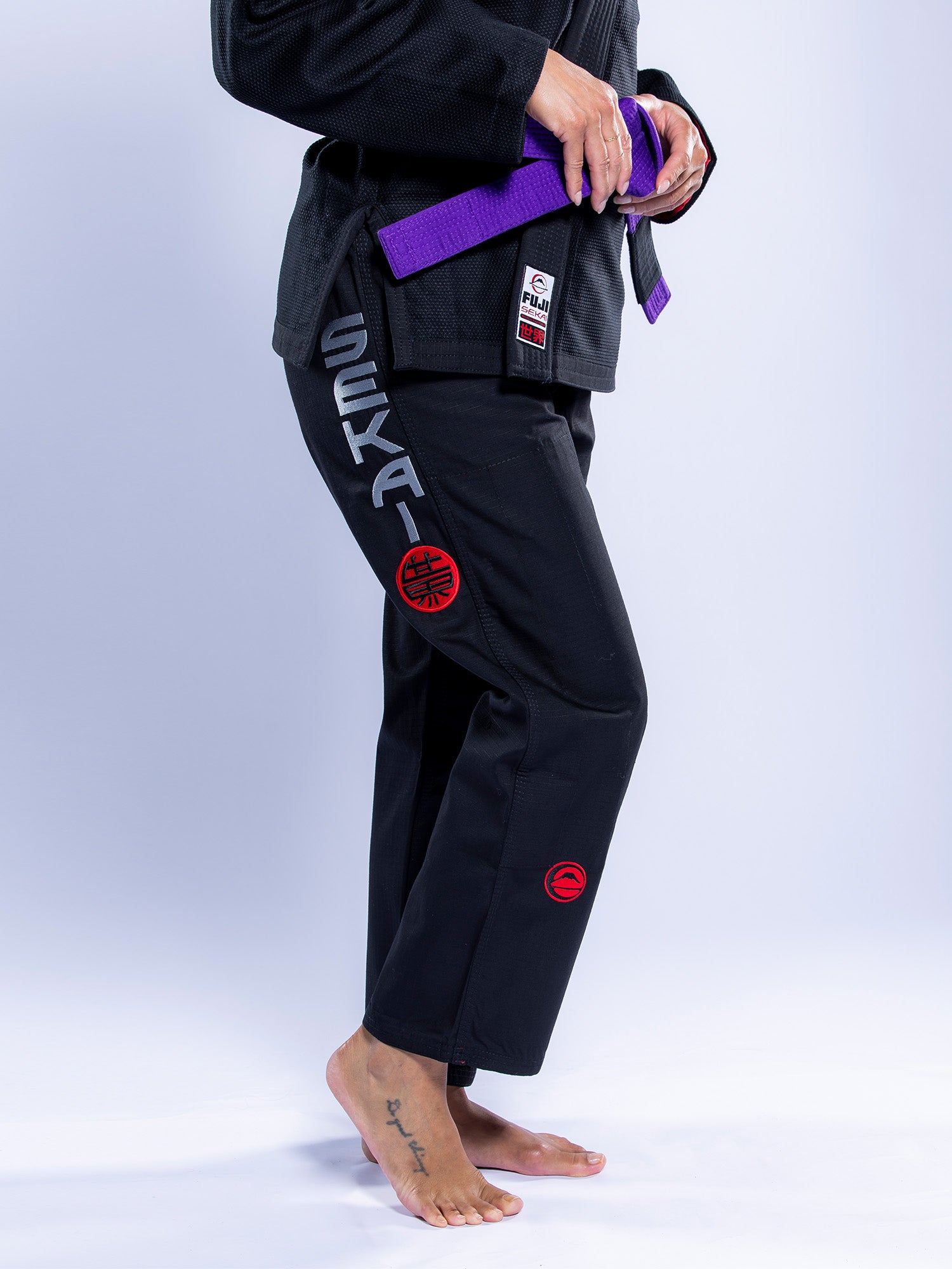 A person stands barefoot wearing the Womens SEKAI 3.0 BJJ Gi Black Grey with red patches and "SEKAI" printed on the pants, a purple belt around their waist, and a small tattoo on their left foot.