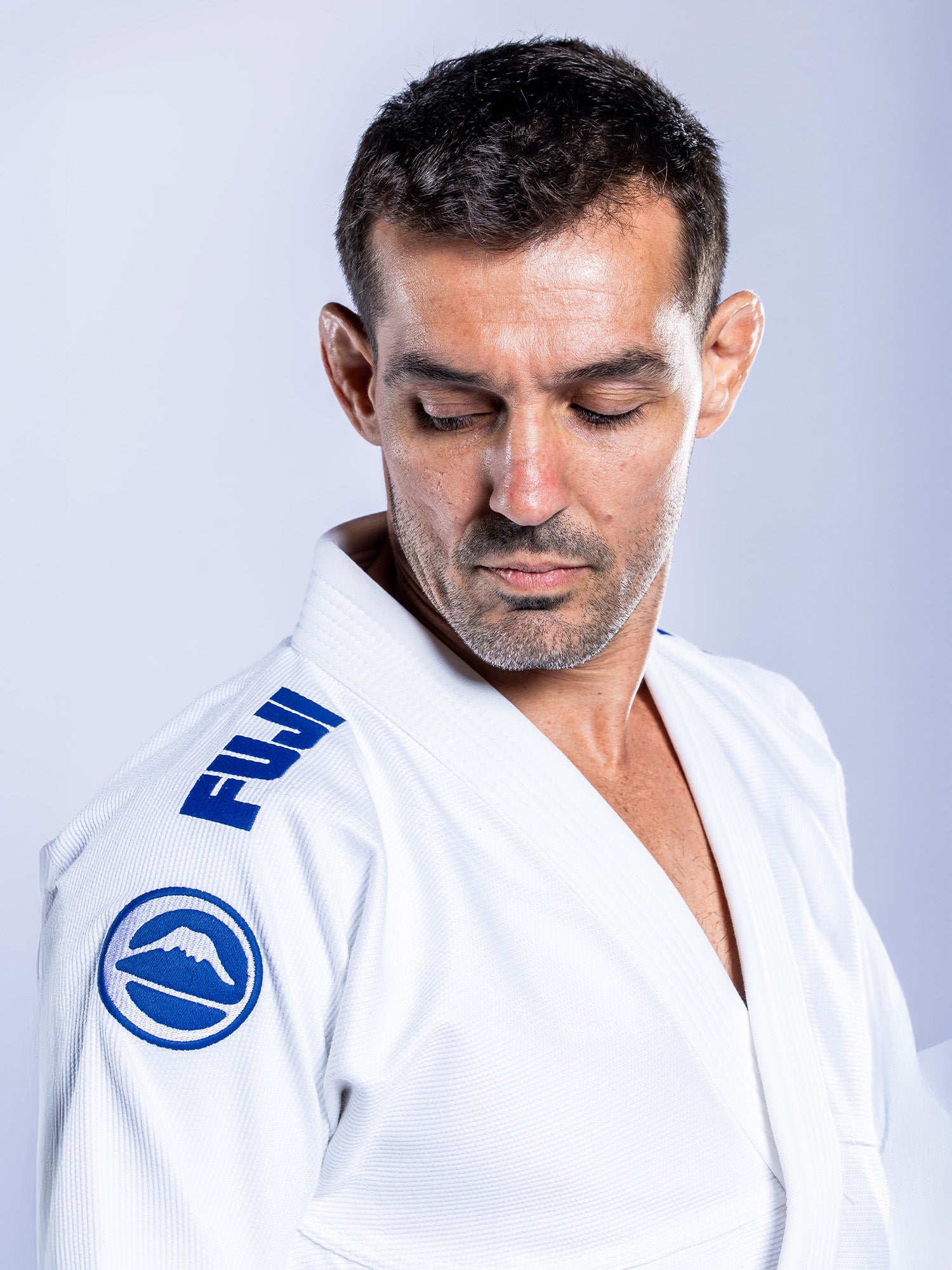 A man wears the Baseline 2.0 BJJ Gi White Navy, looking down over his left shoulder against a plain light background.