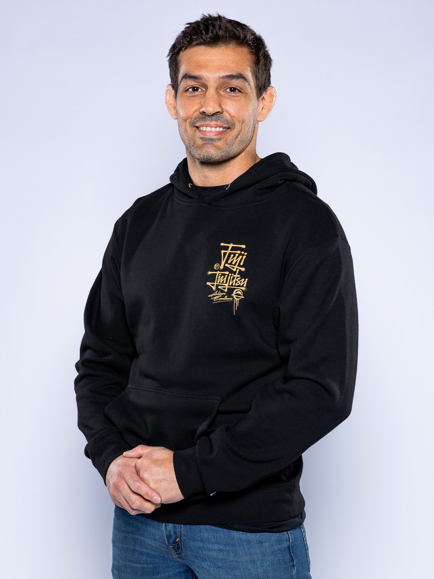 A man with short dark hair smiles against a light background, wearing the Japanese Street Art Hoodie Black with a gold graphic design and blue jeans, his hands loosely clasped in front of him.