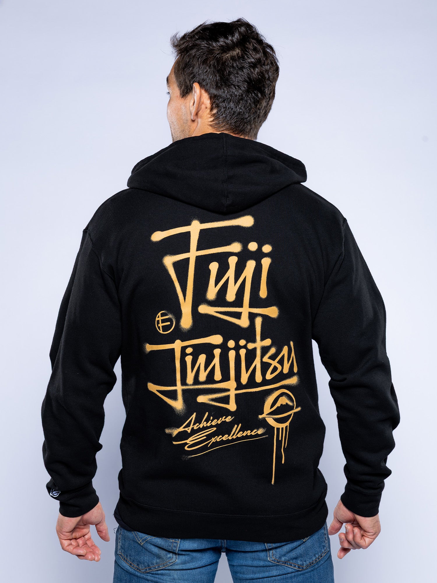 A person with their back to the camera wears the Japanese Street Art Hoodie Black, featuring gold graffiti-style Fuji Jiu Jitsu and Achieve Excellence text. The hood is up, and they’re also wearing blue jeans.