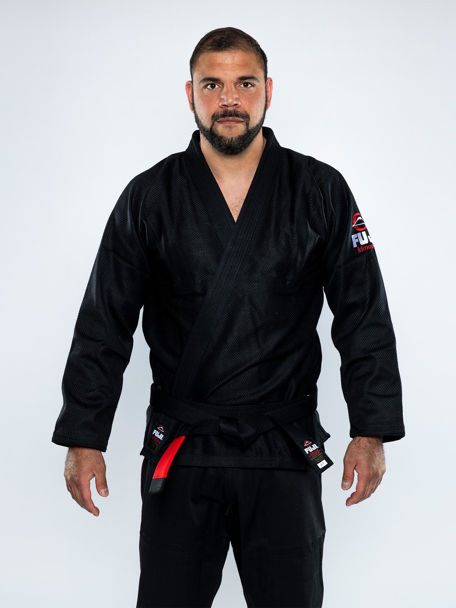 A man with short hair and a beard stands facing forward, wearing the All Around BJJ Gi Black with a patch on the sleeve and a black belt. The background is plain white.