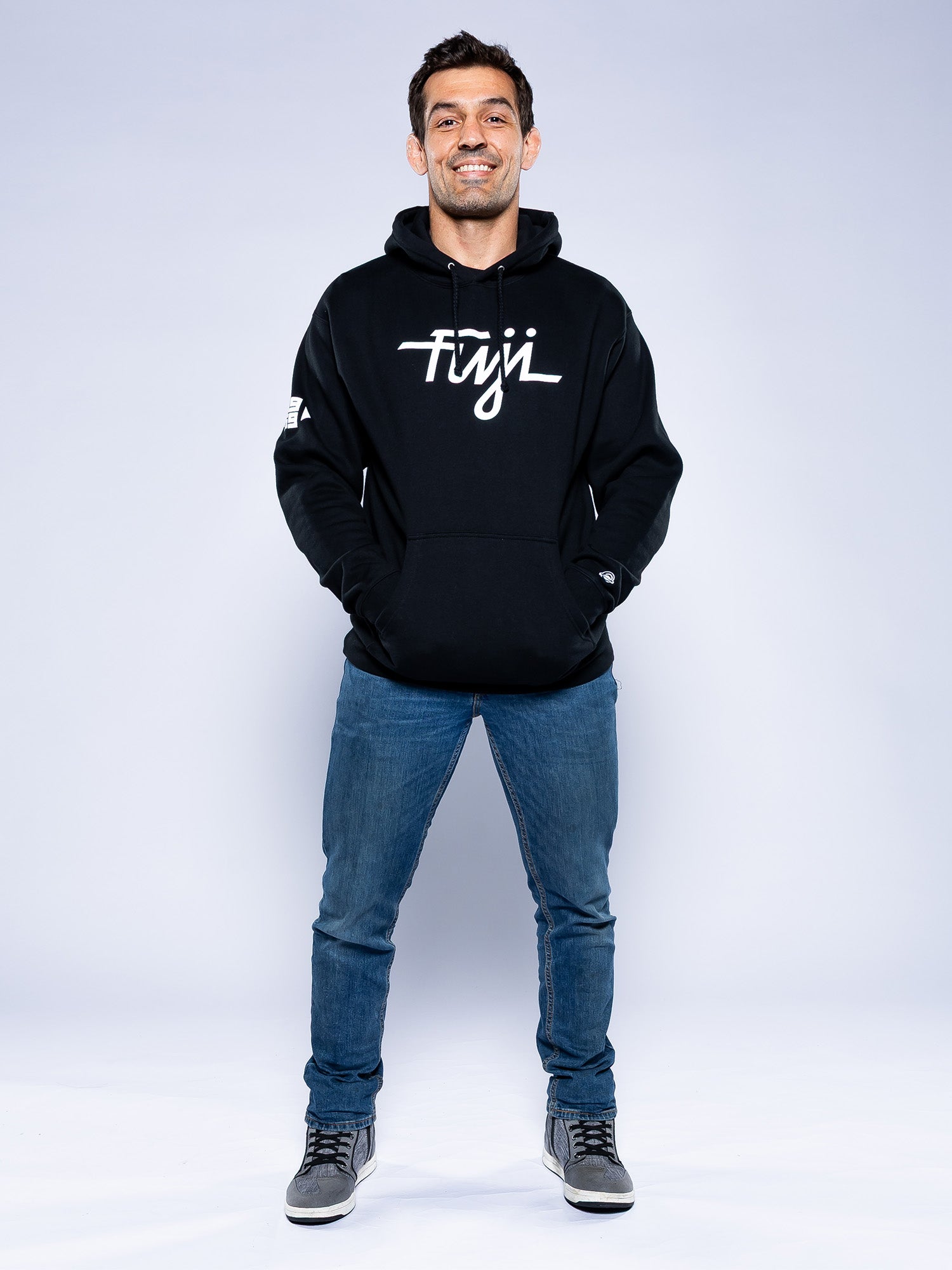A man smiles against a plain light background, wearing the Midnight Collegiate Pull Over Hoodie Black with his hands in the pockets, along with blue jeans and gray sneakers.